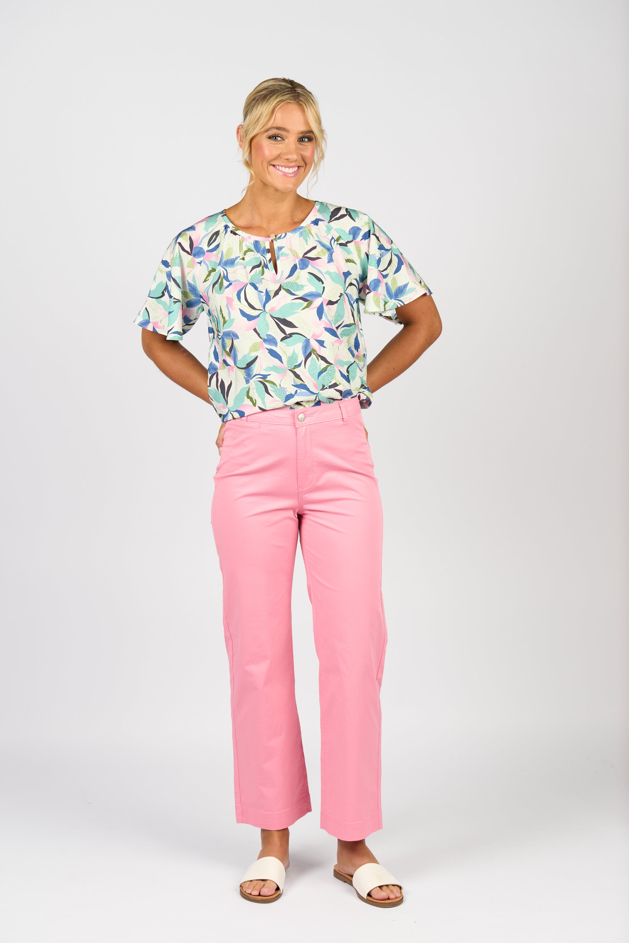 A woman smiles with hands behind her back, wearing a colorful patterned blouse, Vassalli Wide Leg Jean - Jam, white sandals, and a watch against a plain white background.