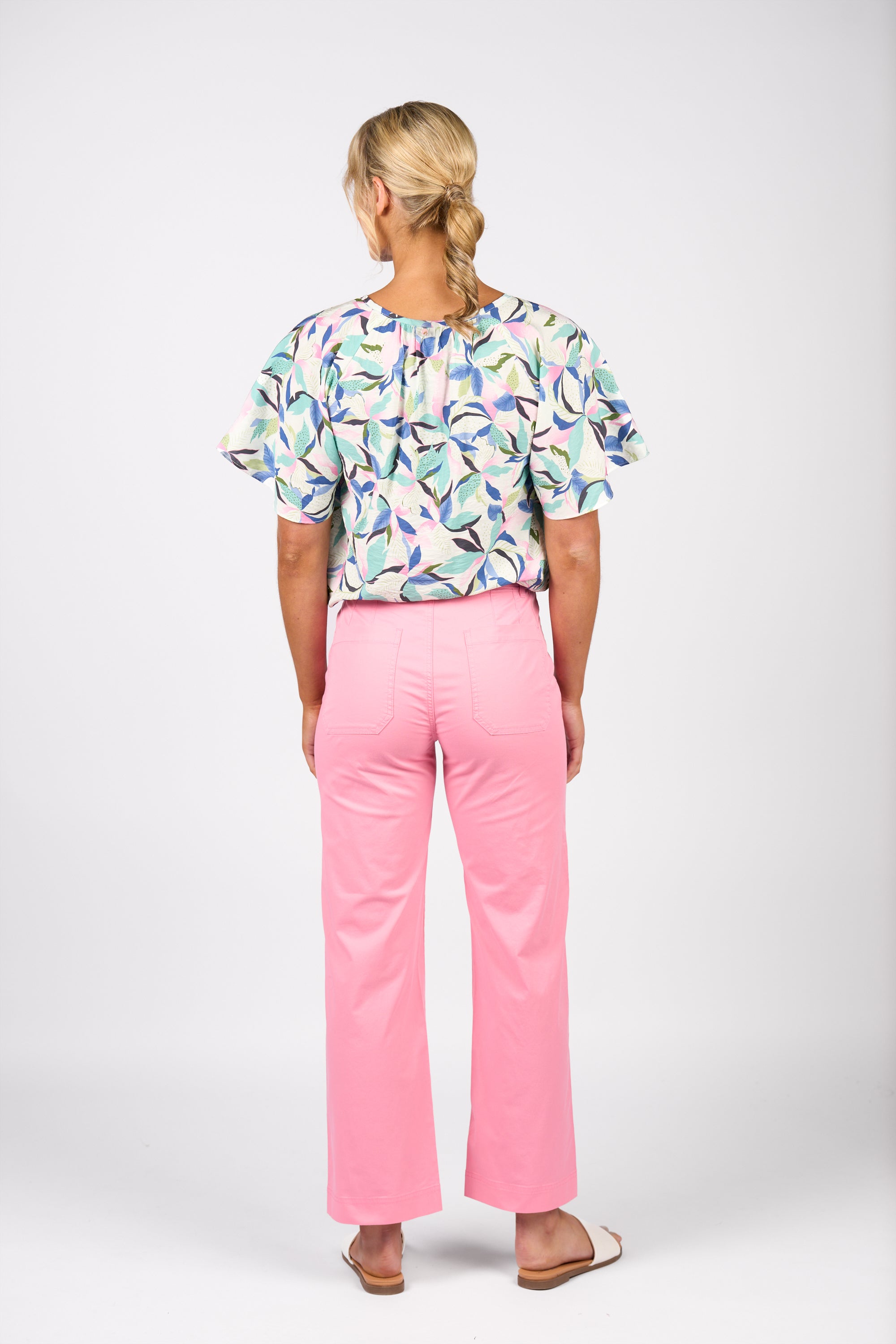 A woman with blonde braided hair faces away, wearing a colorful ruffled blouse, Vassalli’s Wide Leg Jean - Jam, and sandals against a plain white background.