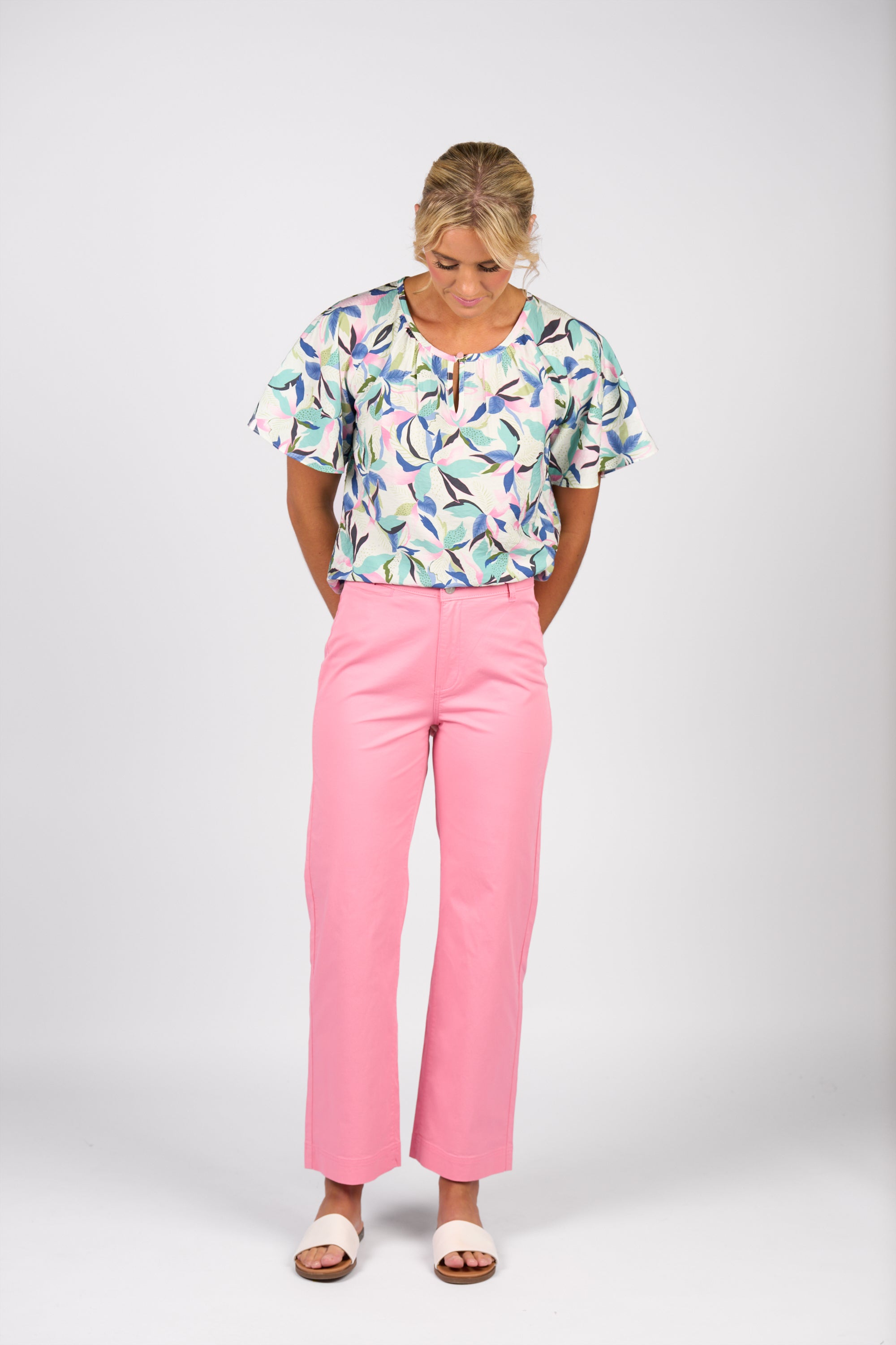 A woman stands against a plain white background, looking down with hands behind her back. She wears a patterned short-sleeve blouse, Vassalli Wide Leg Jean - Jam, and white sandals.