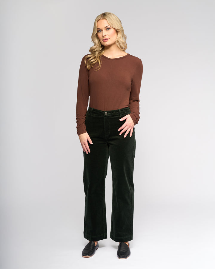 A woman with long blonde hair stands facing forward against a light gray background, wearing Vassalli’s Market Cord Pant in Forest and a brown long-sleeve top, paired with black shoes.