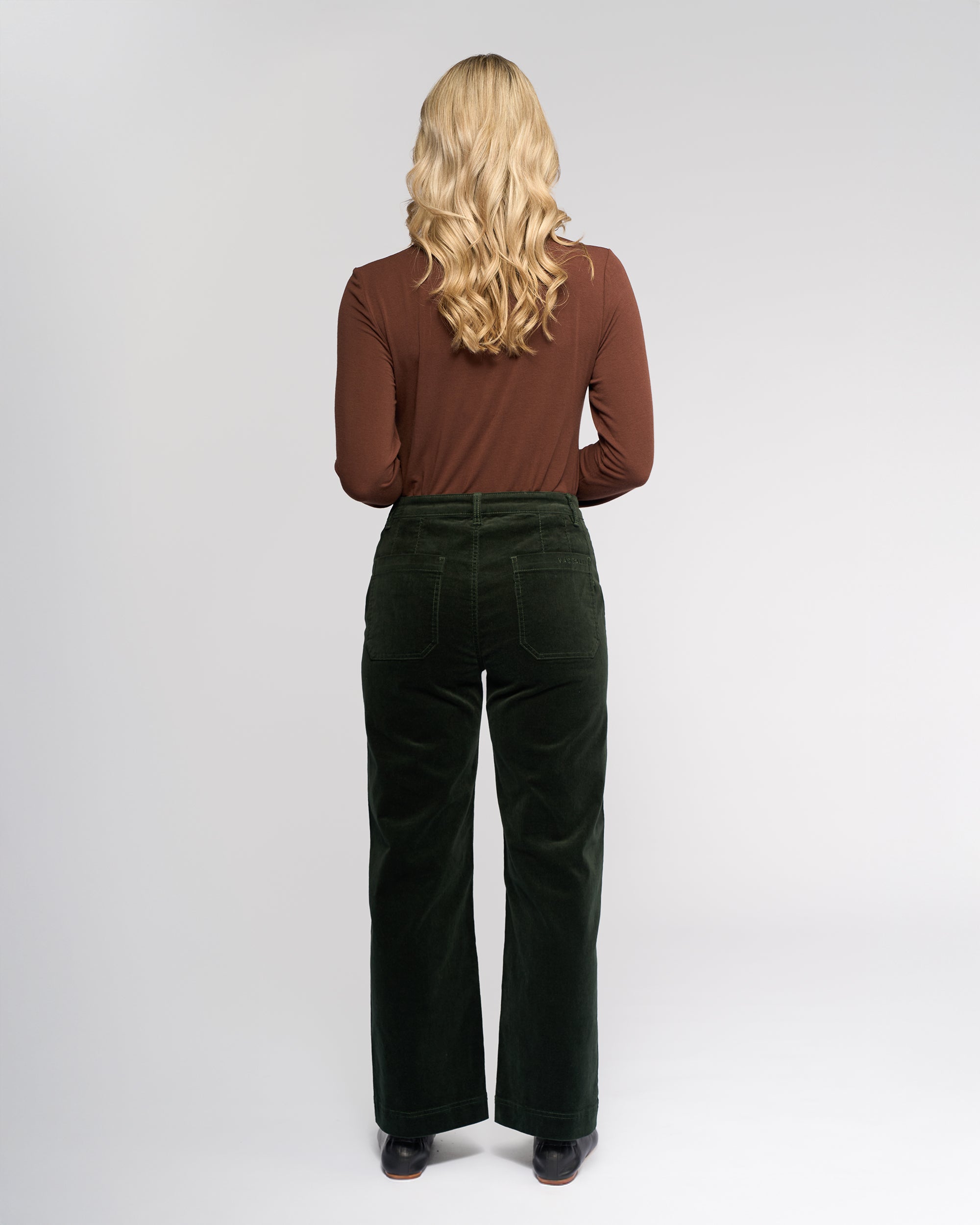 A woman with long blonde hair faces away, wearing a brown long-sleeve shirt and Vassalli Market Cord Pant in Forest against a plain light background.