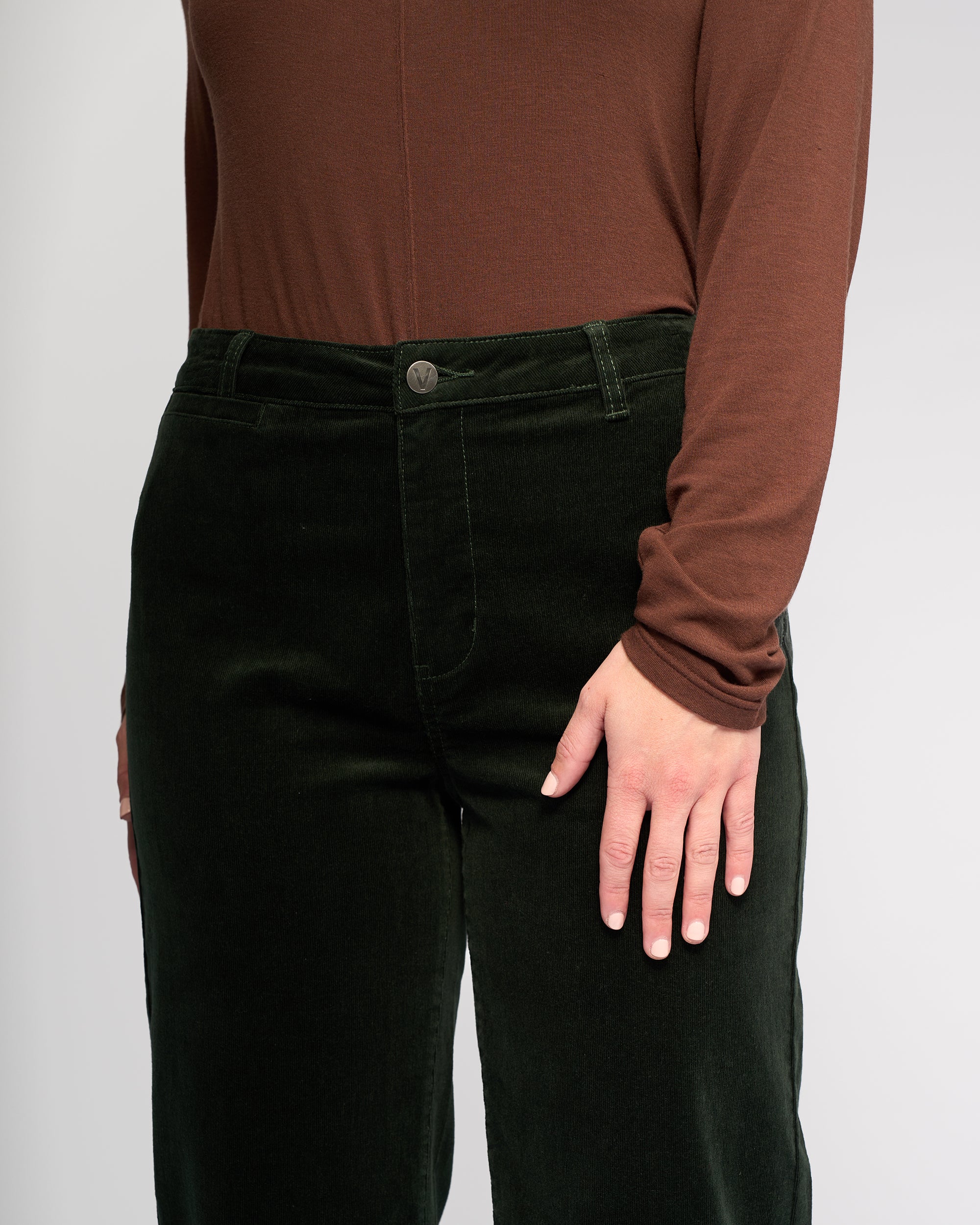 Model wears Vassalli's Market Cord Pant in Forest, a stretch pinwale cord straight-leg ankle-grazer, paired with a long-sleeve brown top. Shown from shoulders to mid-thigh against a plain background, one arm relaxed at their side.