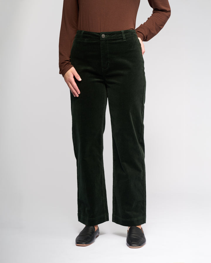 A person wears the Vassalli Market Cord Pant in Forest with a brown long-sleeve top and black slip-on shoes, standing with one hand in the pocket against a plain light gray background.