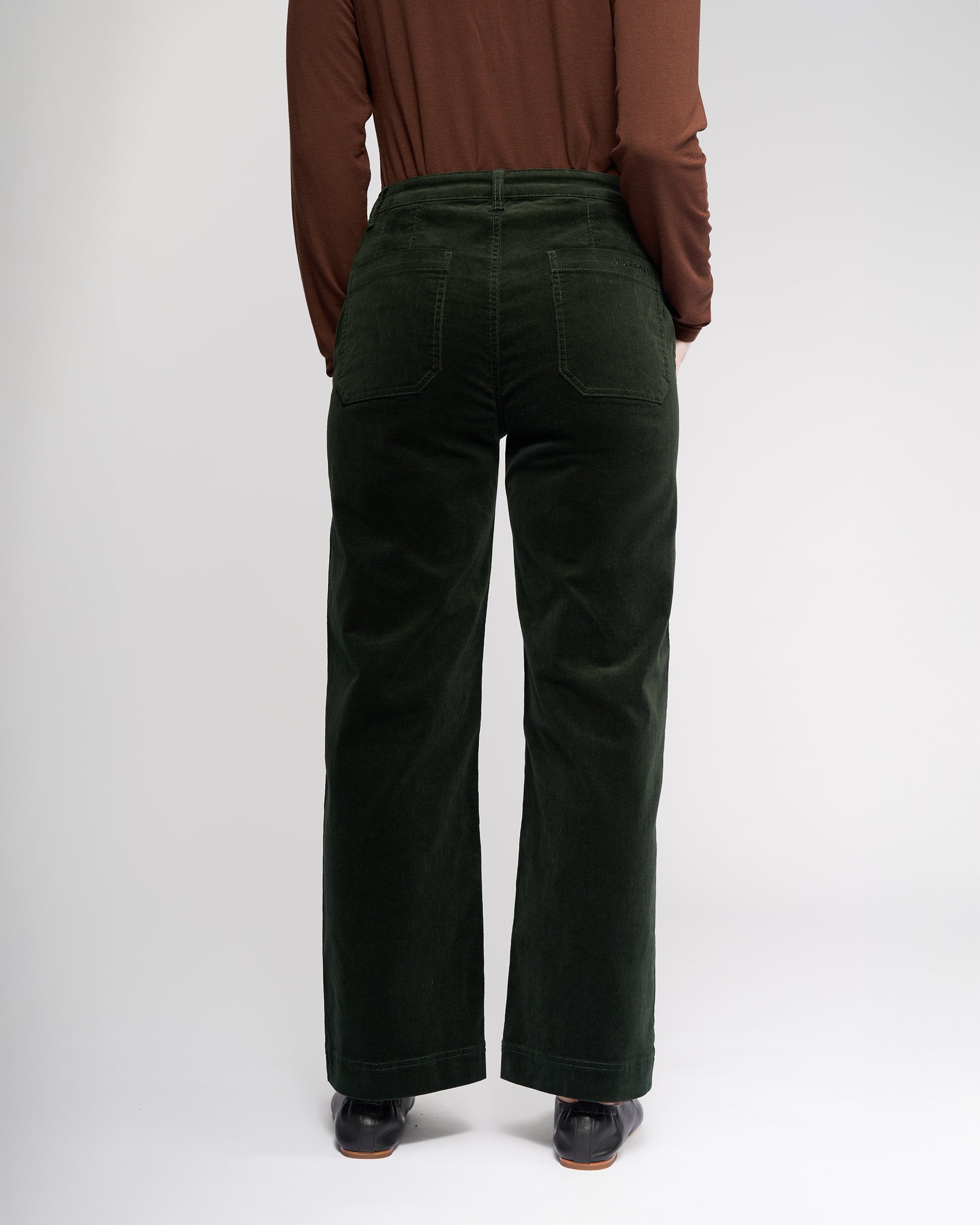 A person faces away from the camera, wearing a long-sleeve brown top and Vassalli Market Cord Pant in Forest—stretch pinwale cord, straight-leg, ankle-grazer fit with back pockets—and dark shoes. The plain background is light.