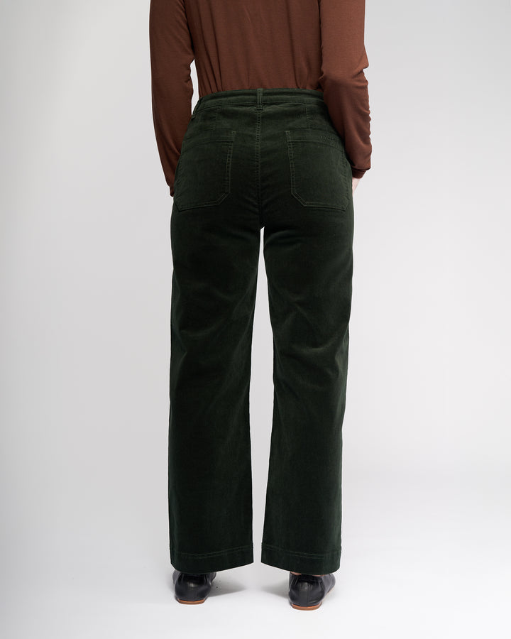 A person faces away from the camera, wearing a long-sleeve brown top and Vassalli Market Cord Pant in Forest—stretch pinwale cord, straight-leg, ankle-grazer fit with back pockets—and dark shoes. The plain background is light.