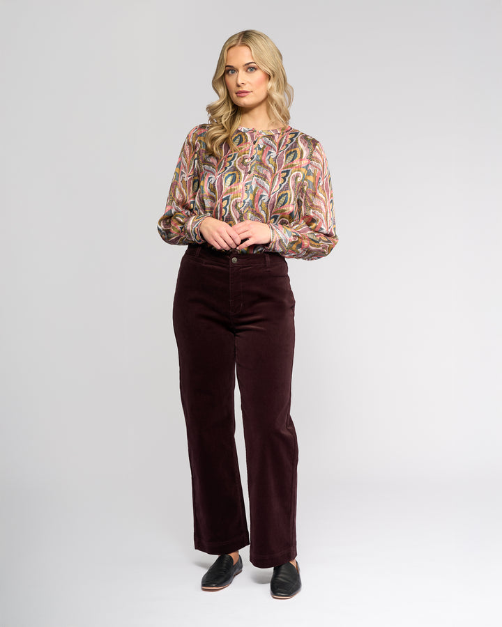 A woman with long blonde hair stands facing forward in a colorful patterned blouse, Vassalli's Market Cord Pant - Merlot, and black slip-on shoes against a plain light background.