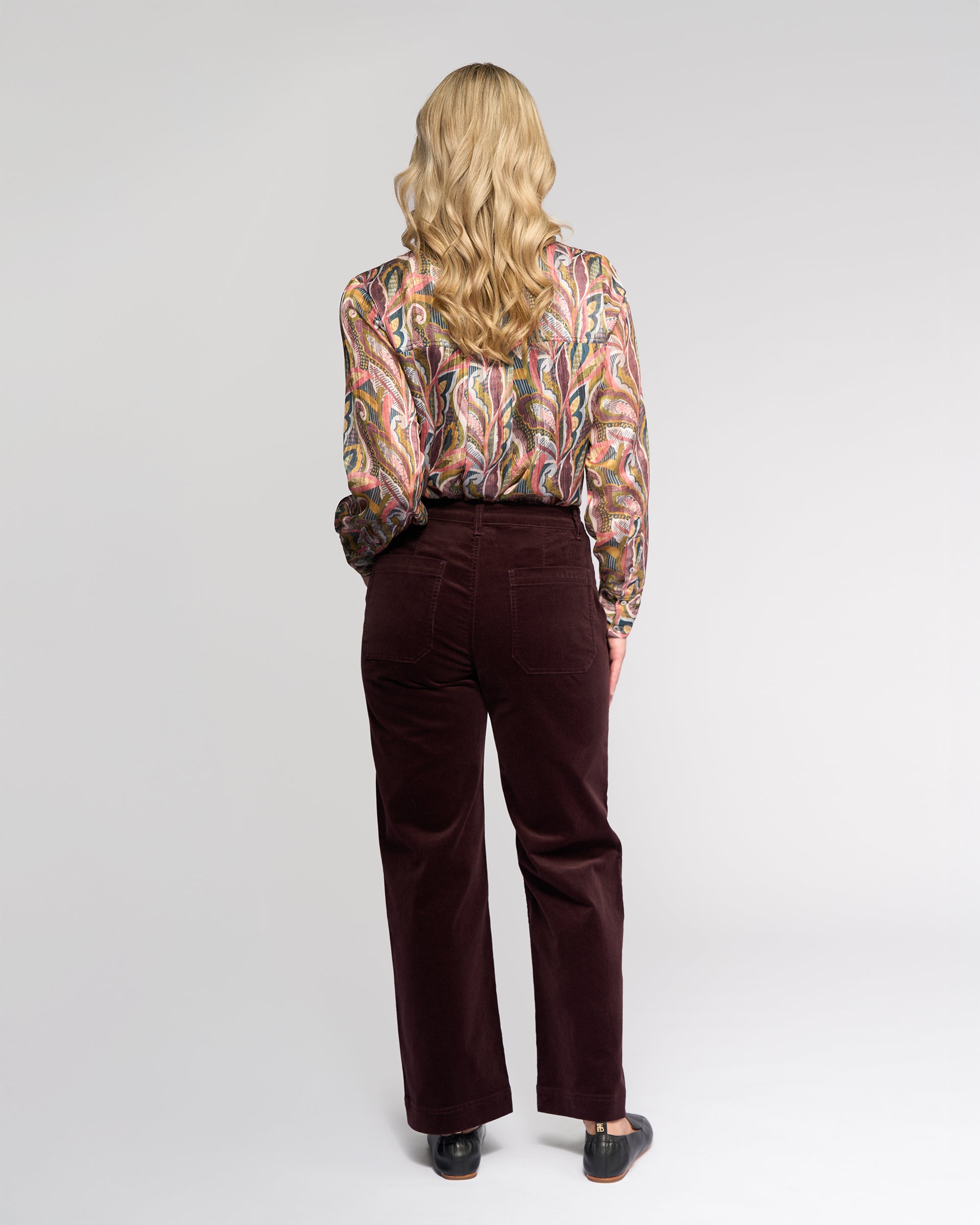 A person with long, wavy blonde hair faces away, wearing a colorful paisley blouse, Vassalli Market Cord Pant in Merlot, and black shoes against a plain light background.