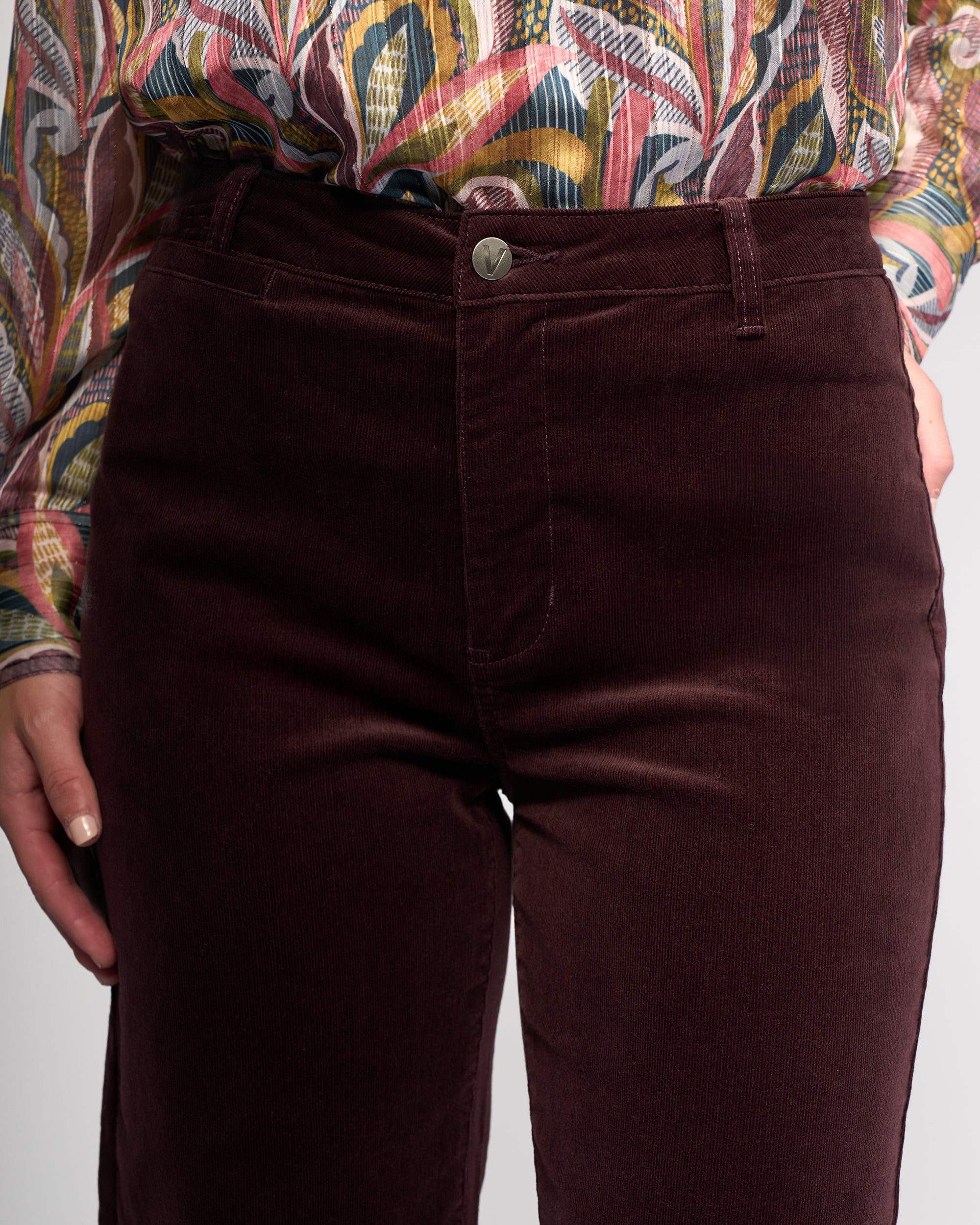 A person wears Vassalli's Market Cord Pant in Merlot with a colorful patterned shirt, one hand by their side. The image highlights the waist and upper legs.