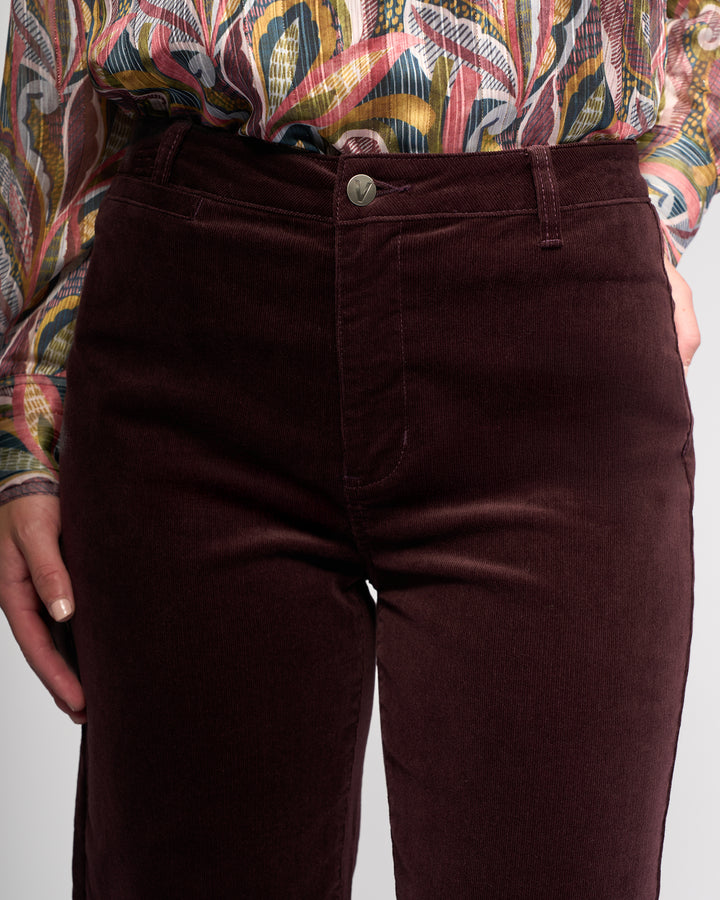 A person wears Vassalli's Market Cord Pant in Merlot with a colorful patterned shirt, one hand by their side. The image highlights the waist and upper legs.