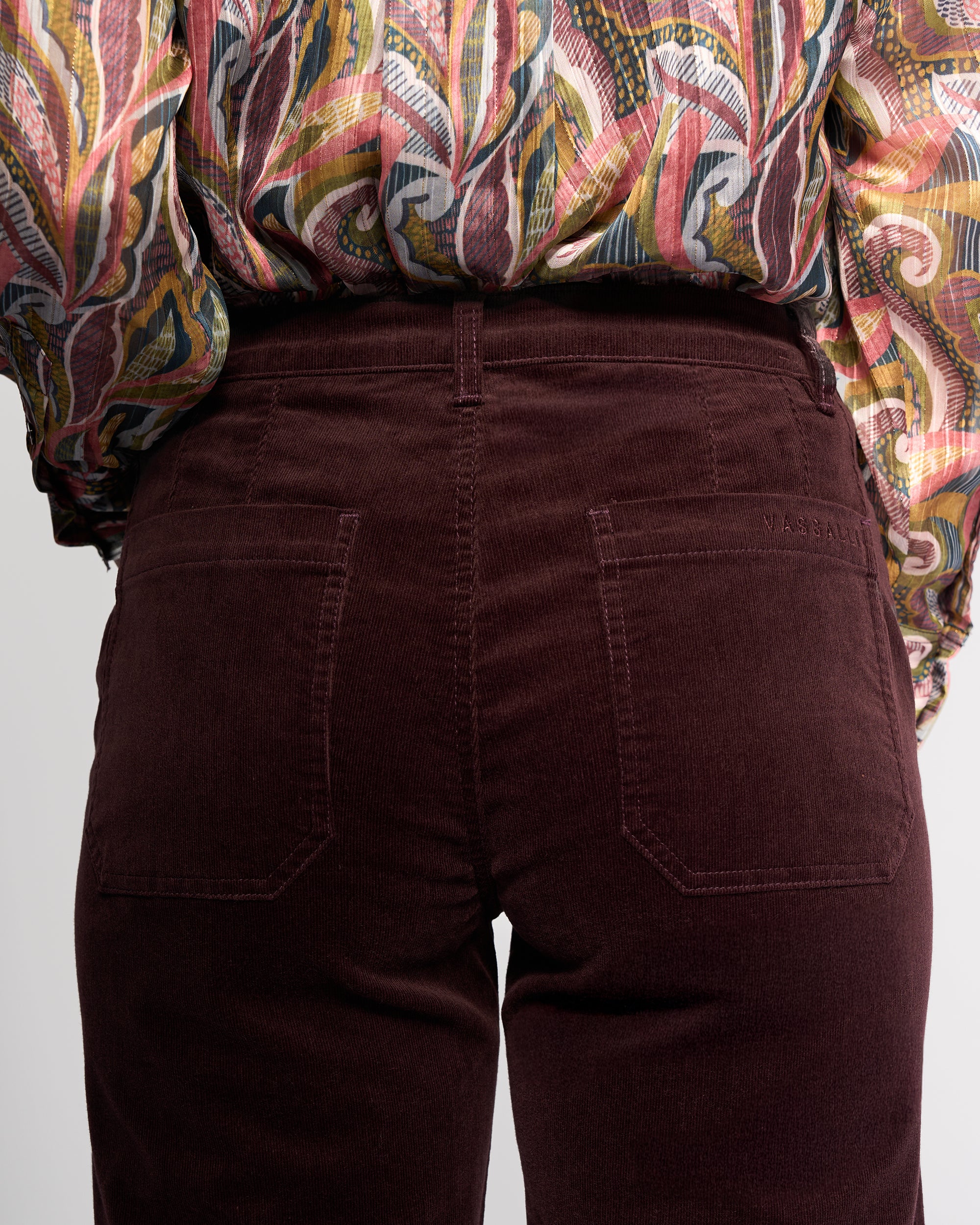 A close-up of a person wearing Vassalli's Market Cord Pant in Merlot, shown from the waist down with both back pockets visible, paired with a colorful patterned shirt.