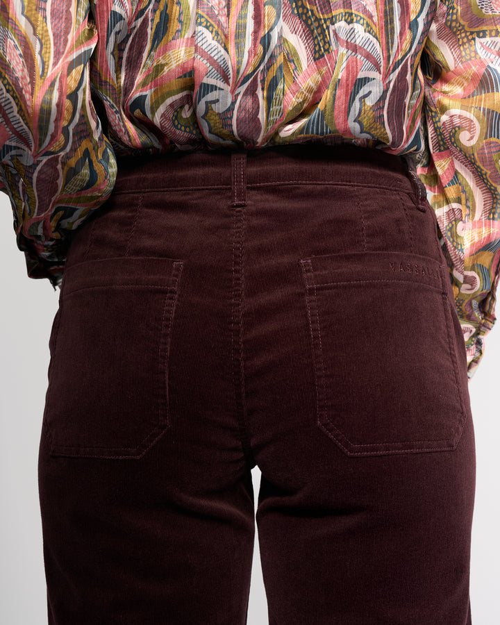 A close-up of a person wearing Vassalli's Market Cord Pant in Merlot, shown from the waist down with both back pockets visible, paired with a colorful patterned shirt.
