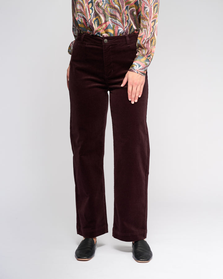 Against a plain background, a person (head out of frame) wears Vassalli's Market Cord Pant in Merlot, a colorful patterned long-sleeve shirt, and black slip-on shoes. Their left hand is on their hip.