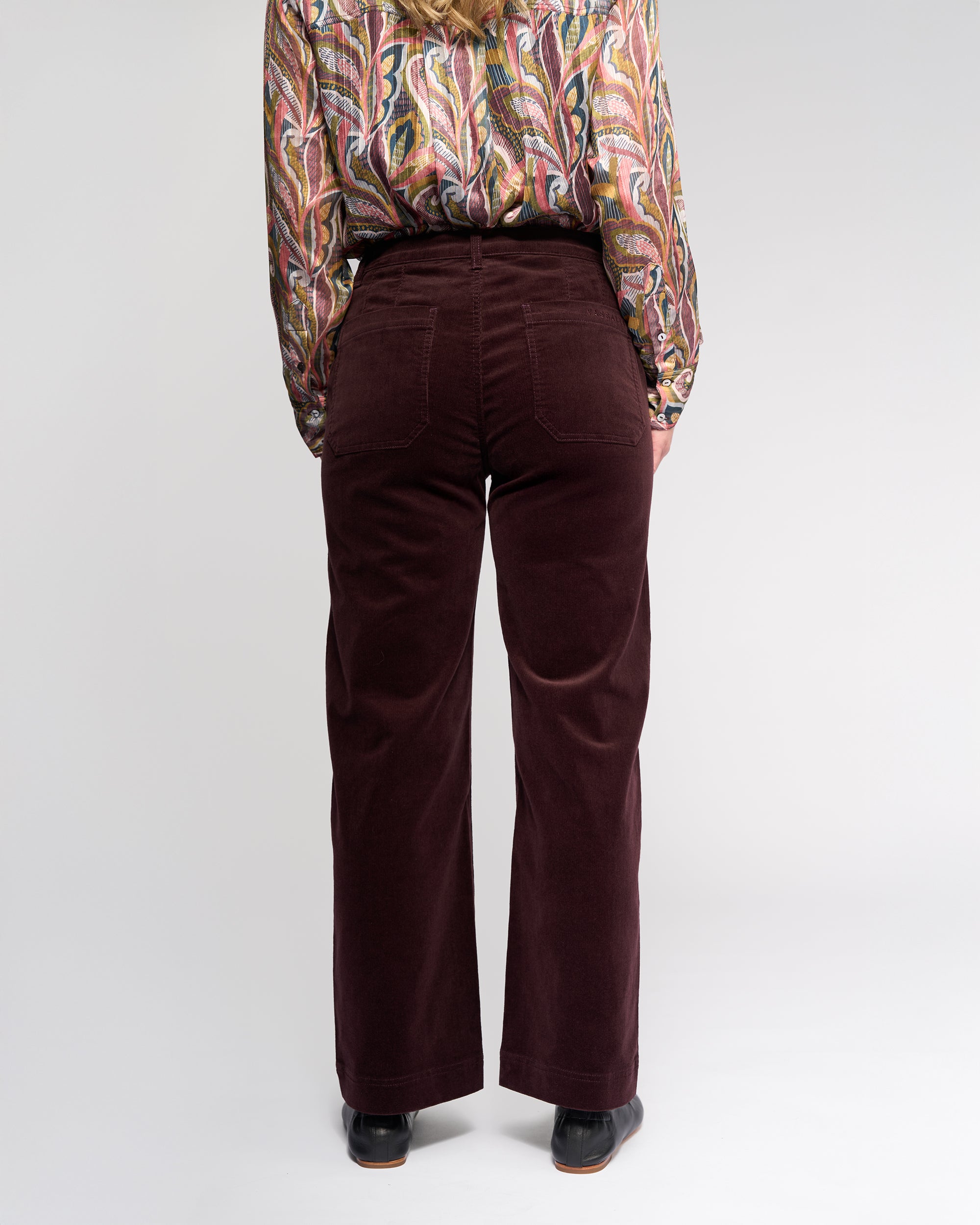 A person stands facing away, wearing Vassalli's Market Cord Pant in Merlot and a colorful paisley long-sleeve shirt with black shoes. The plain light background highlights the maroon accents of the pants.