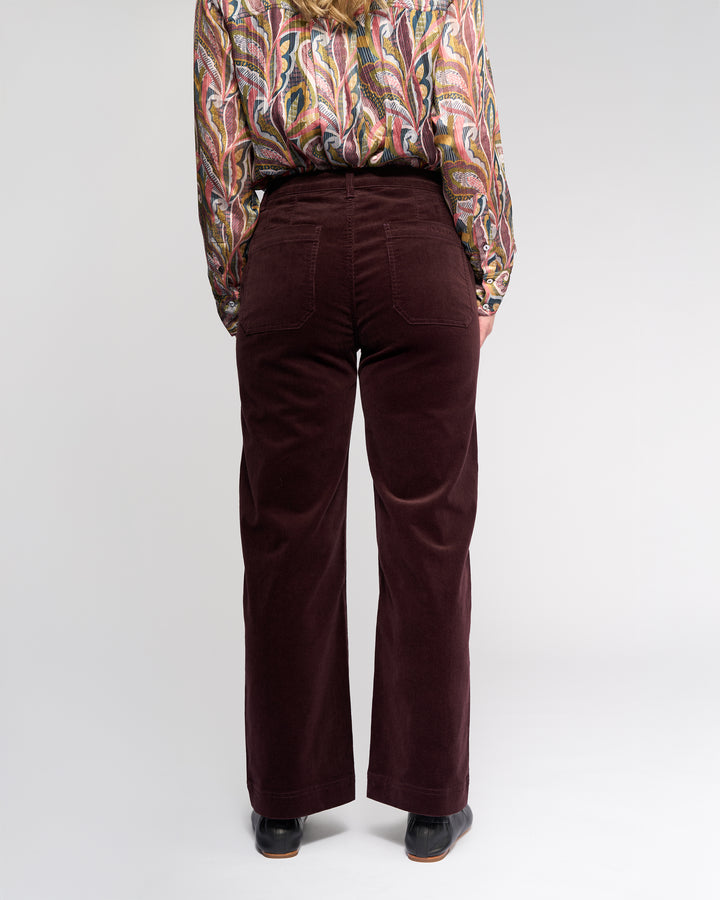 A person stands facing away, wearing Vassalli's Market Cord Pant in Merlot and a colorful paisley long-sleeve shirt with black shoes. The plain light background highlights the maroon accents of the pants.