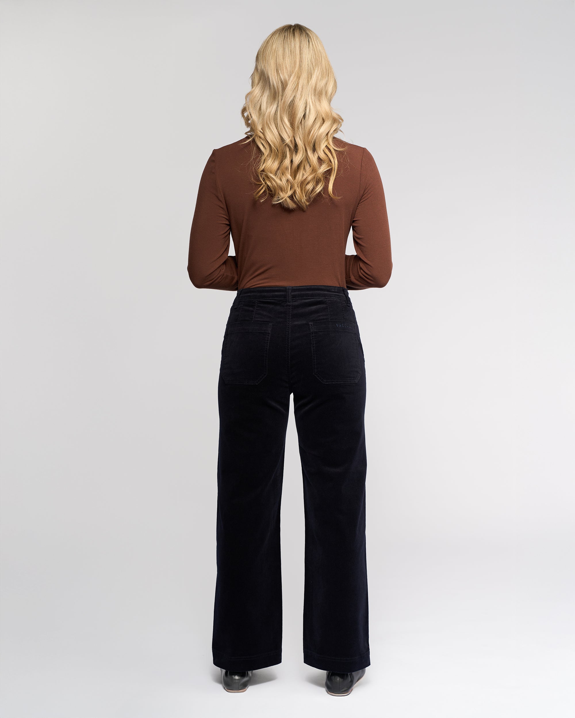 A person with long, wavy blonde hair faces away from the camera, wearing a brown long-sleeve top and Vassalli’s Market Cord Pant - Midnight against a plain, light background.