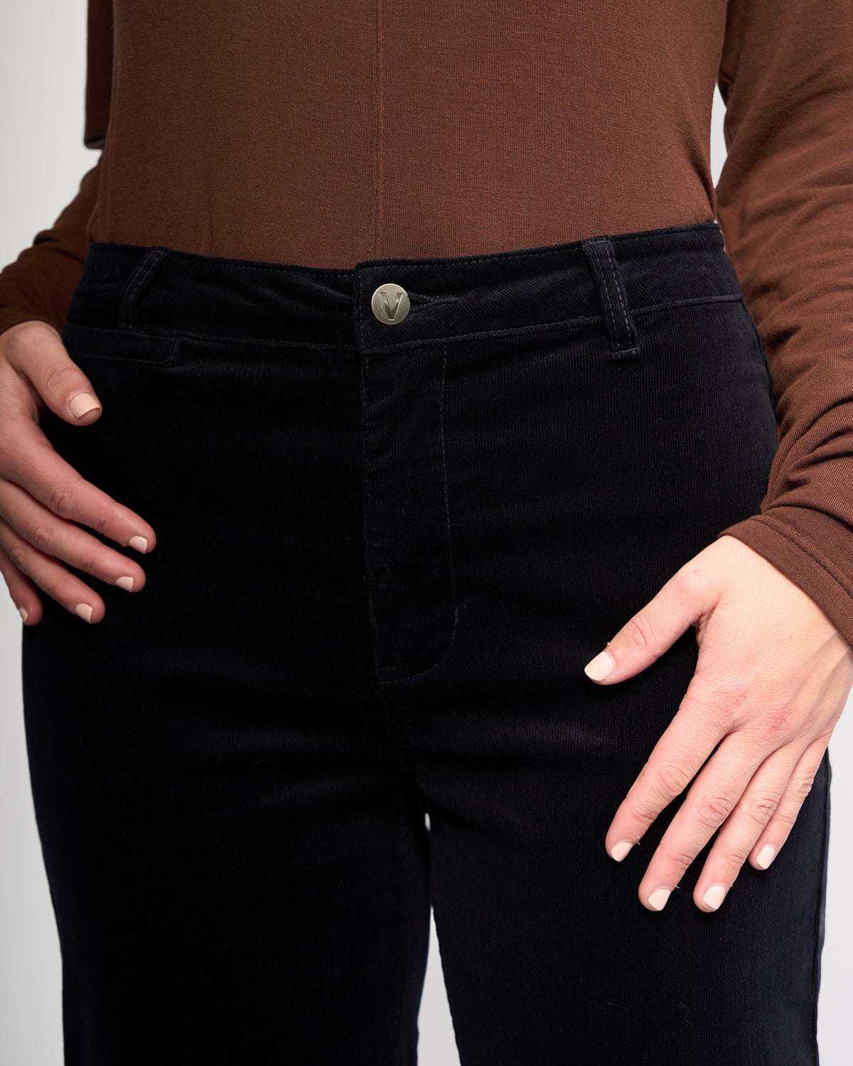 A person models the Vassalli Market Cord Pant in Midnight, paired with a brown long-sleeve top and stretch pinwale cord shirt, standing hands on hips to display the pants' front.