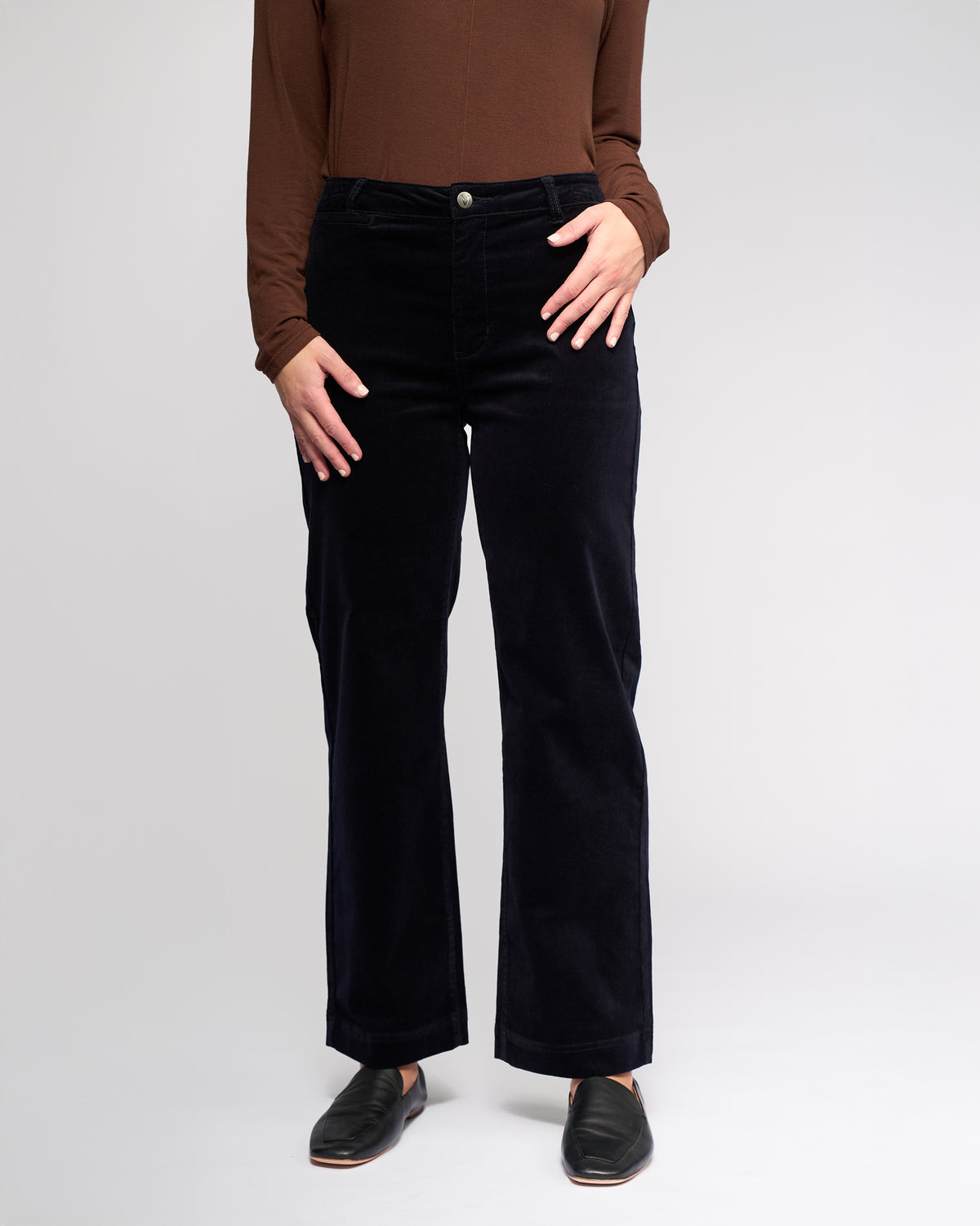 A person wears Vassalli's Market Cord Pant in Midnight, paired with a brown long-sleeve top and black slip-on shoes. Standing against a light backdrop, their hands rest near the pants’ front pockets; their head is not visible.
