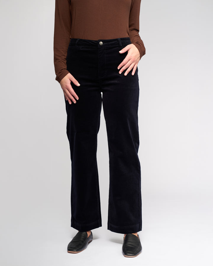 A person wears Vassalli's Market Cord Pant in Midnight, paired with a brown long-sleeve top and black slip-on shoes. Standing against a light backdrop, their hands rest near the pants’ front pockets; their head is not visible.