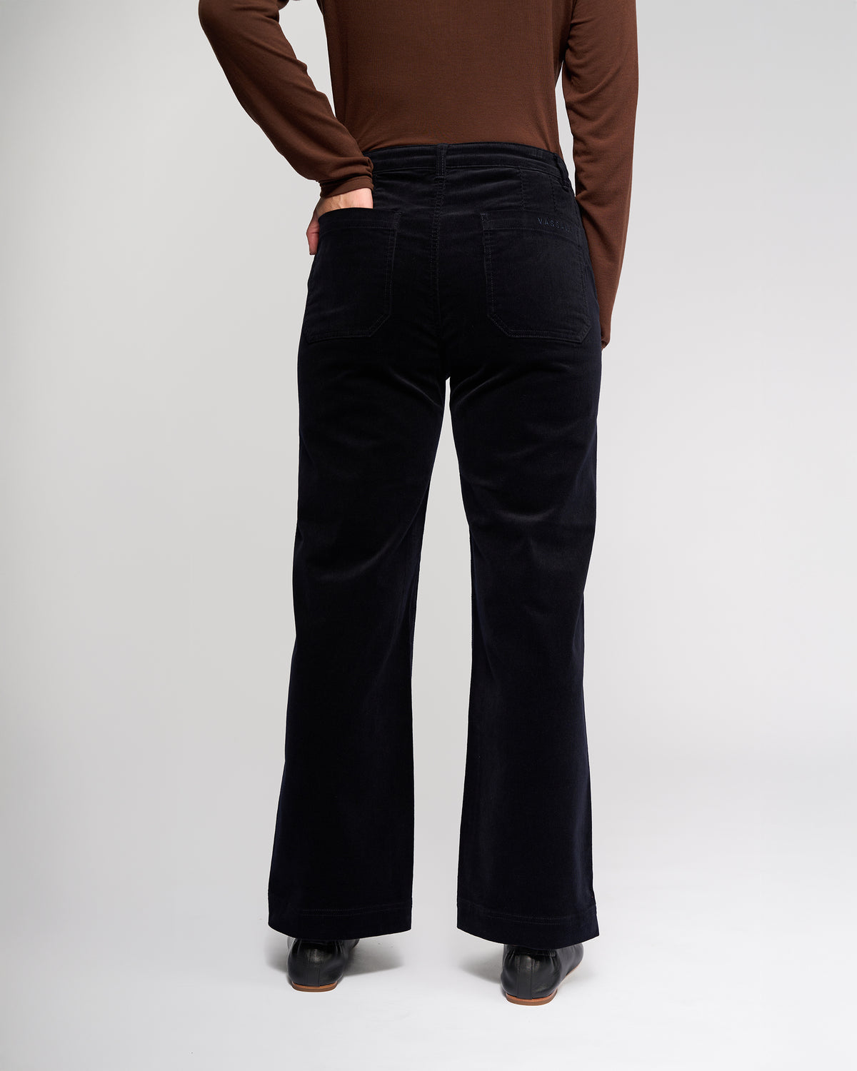 A person wearing a brown long-sleeve top and Vassalli Market Cord Pant in Midnight stands with their back to the camera, one hand on their hip, set against a light plain background.