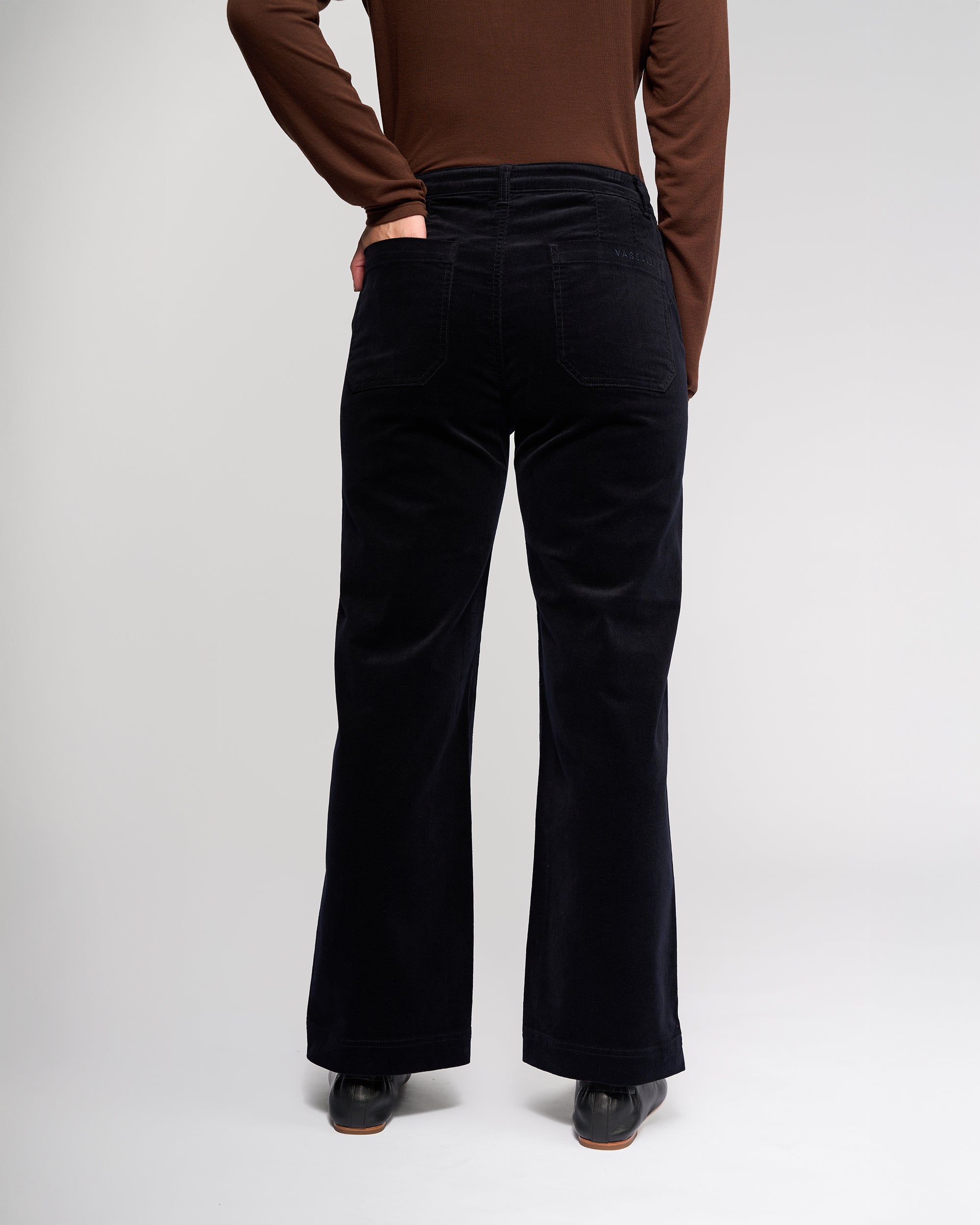 A person wearing a brown long-sleeve top and Vassalli Market Cord Pant in Midnight stands with their back to the camera, one hand on their hip, set against a light plain background.