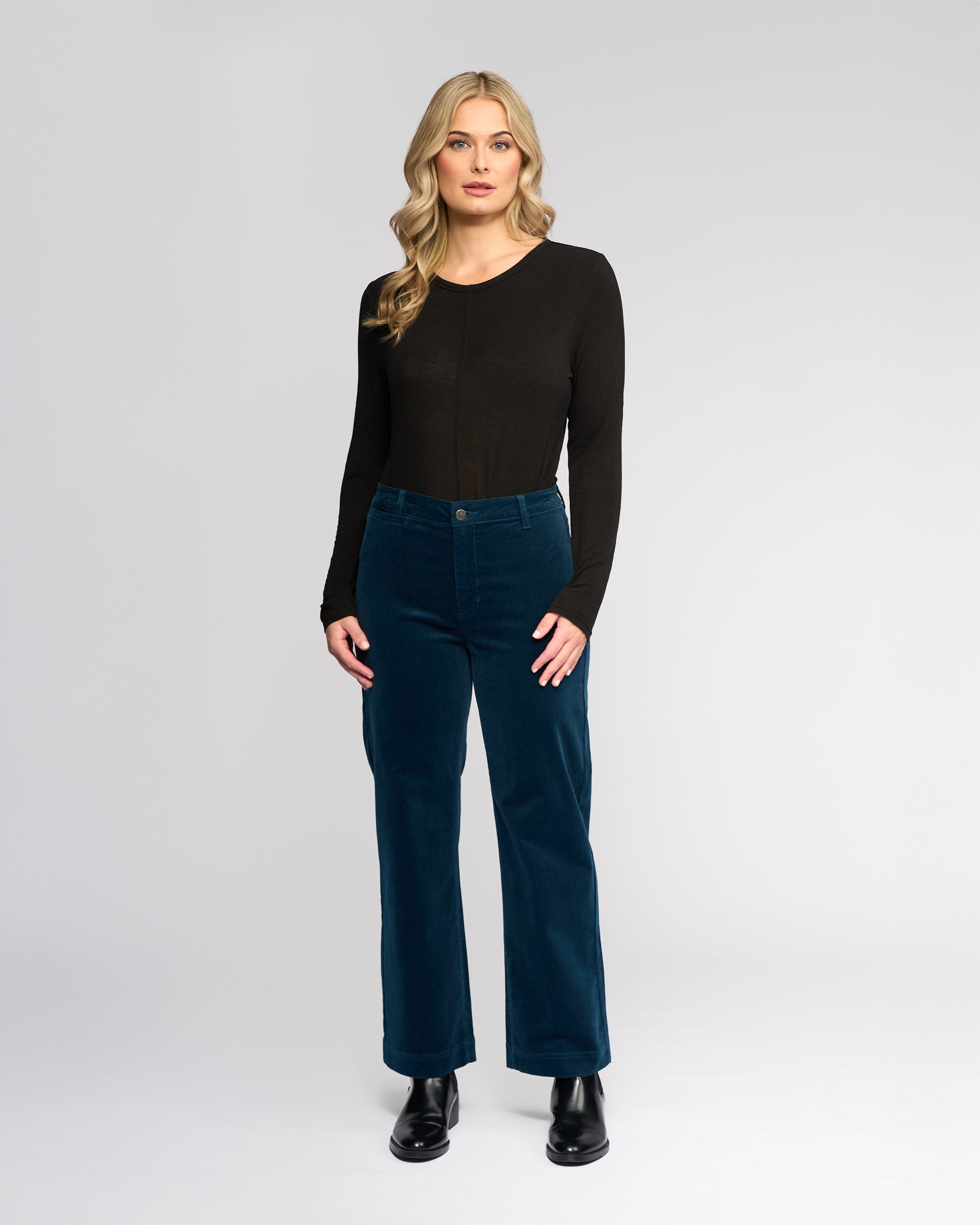 A woman with long blonde hair stands against a plain light background, wearing a Vassalli Market Cord Pant in Petrol, a long-sleeve black top, and black boots. She faces forward with a neutral expression.