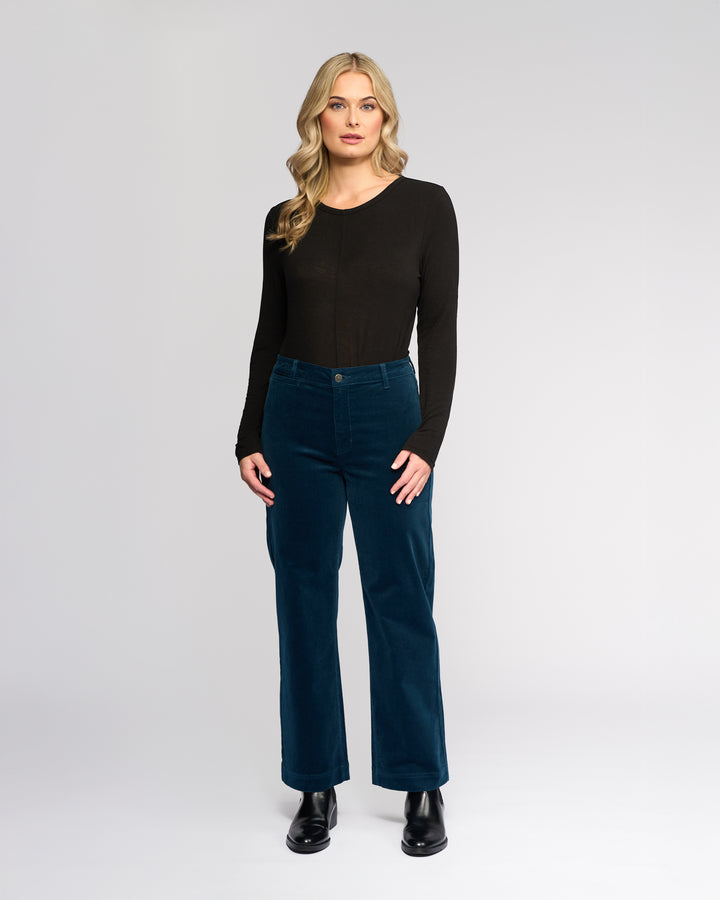 A woman with long blonde hair stands against a plain light background, wearing a Vassalli Market Cord Pant in Petrol, a long-sleeve black top, and black boots. She faces forward with a neutral expression.