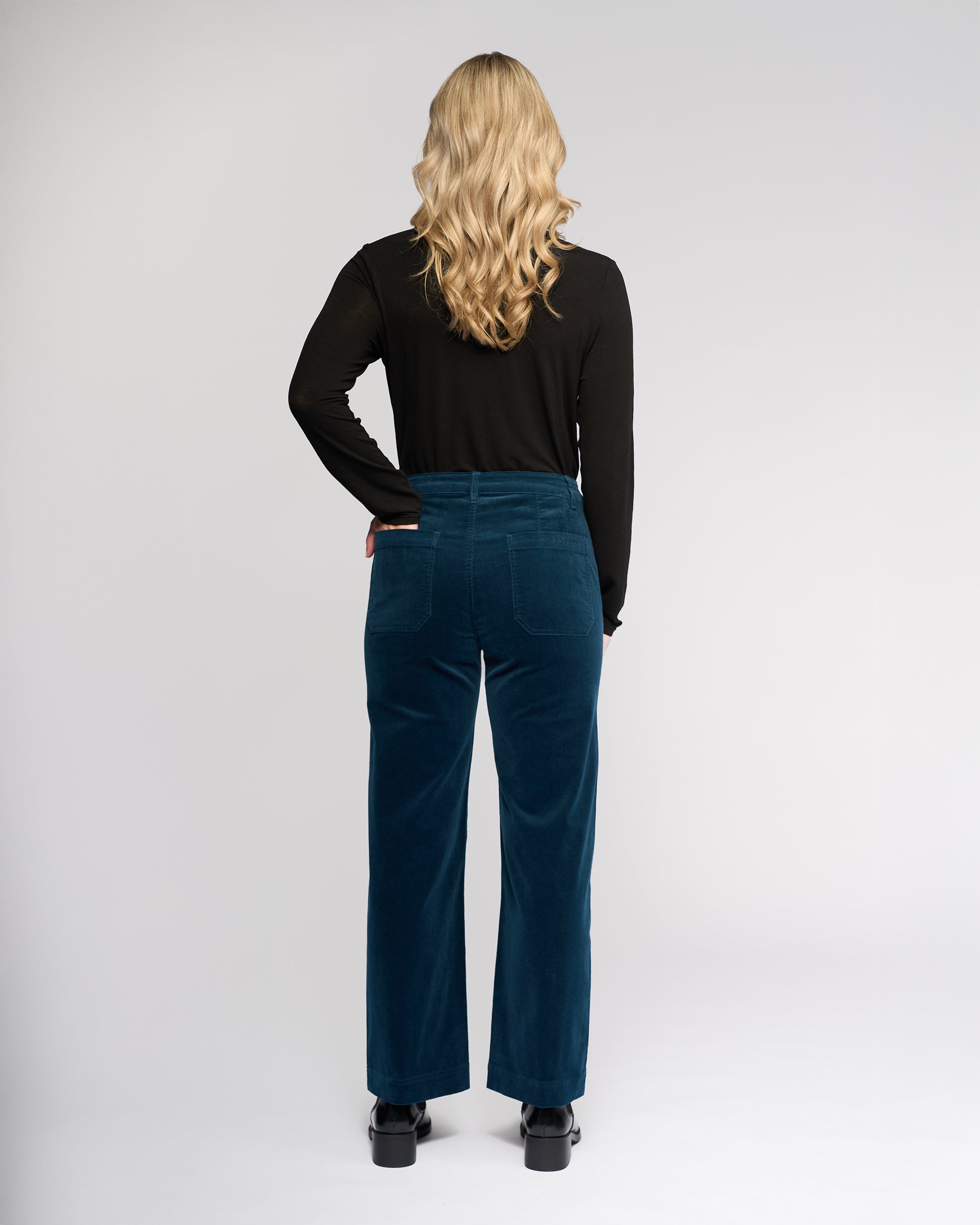A person with long, wavy blonde hair stands facing away in a black long-sleeve top, black shoes, and Vassalli's Market Cord Pant in Petrol against a plain light background.