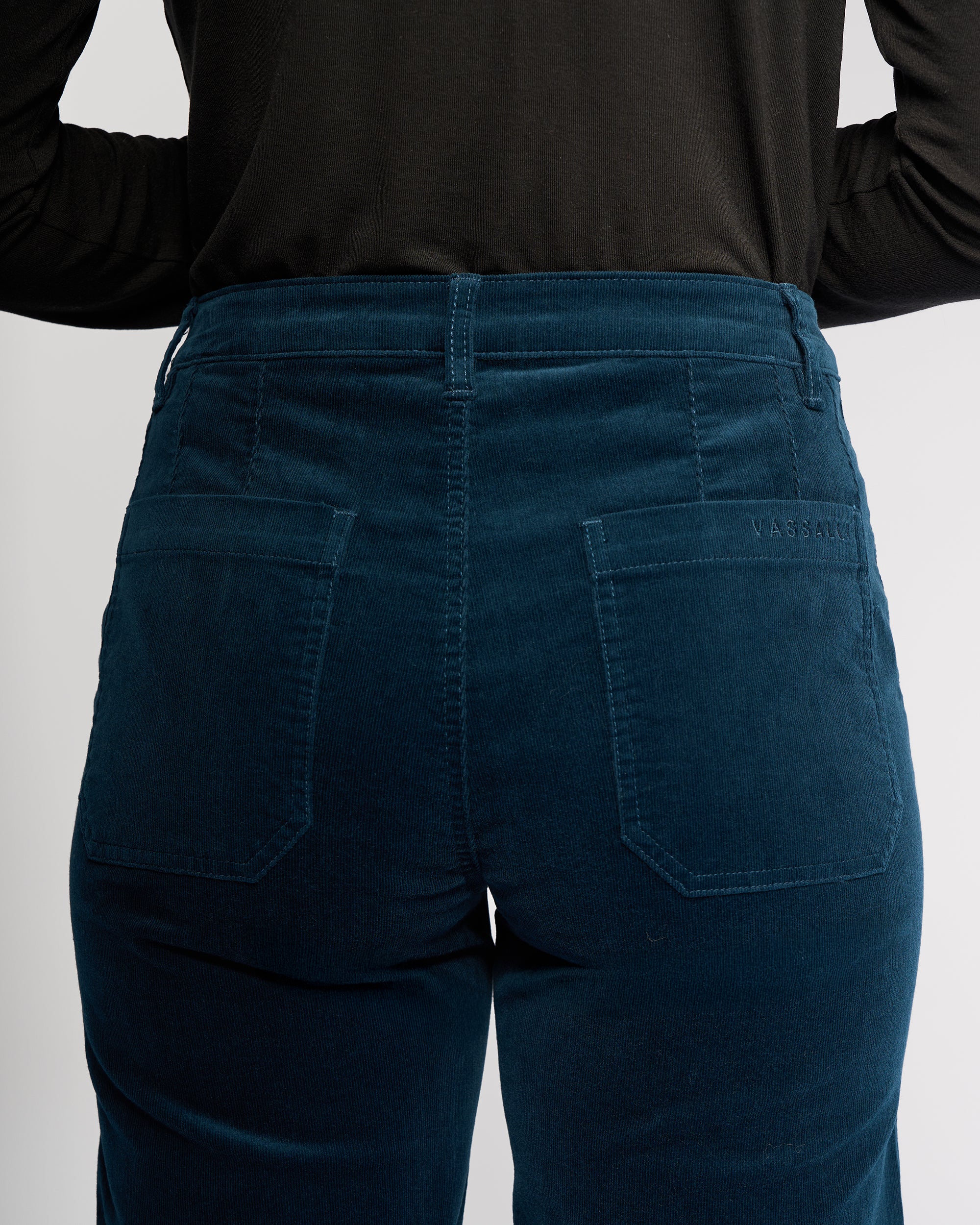 A person wears Vassalli’s Market Cord Pant in Petrol, shown from the waist down with their back to the camera. The straight-leg pants have visible stitching, two back pockets, and an ankle-grazer length, paired with a black top.