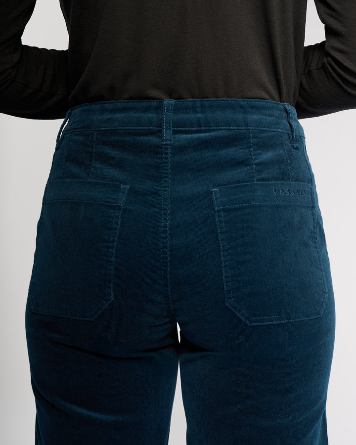 A person wears Vassalli’s Market Cord Pant in Petrol, shown from the waist down with their back to the camera. The straight-leg pants have visible stitching, two back pockets, and an ankle-grazer length, paired with a black top.