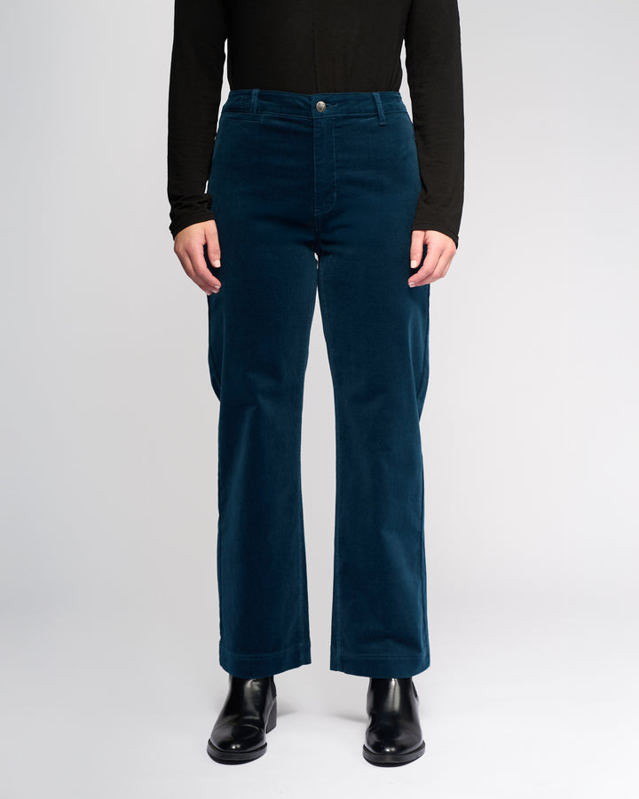A person is shown from the neck down wearing a black long-sleeve top, black boots, and Vassalli’s Market Cord Pant in Petrol, standing against a plain white background.