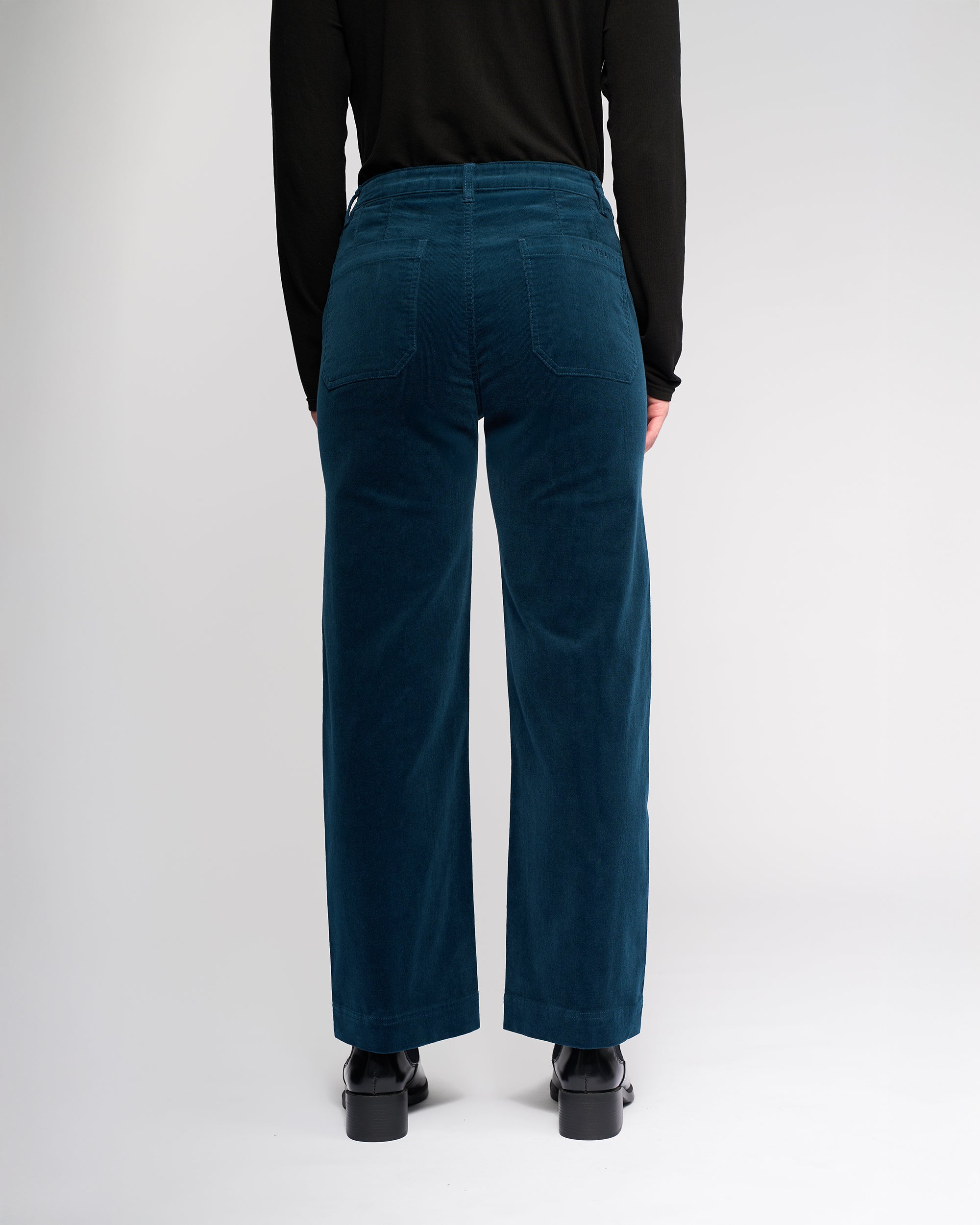 A person stands with their back to the camera, wearing a black long-sleeve top and Vassalli Market Cord Pant in Petrol. The pants feature patch pockets and an ankle-grazer length. The background is plain white.