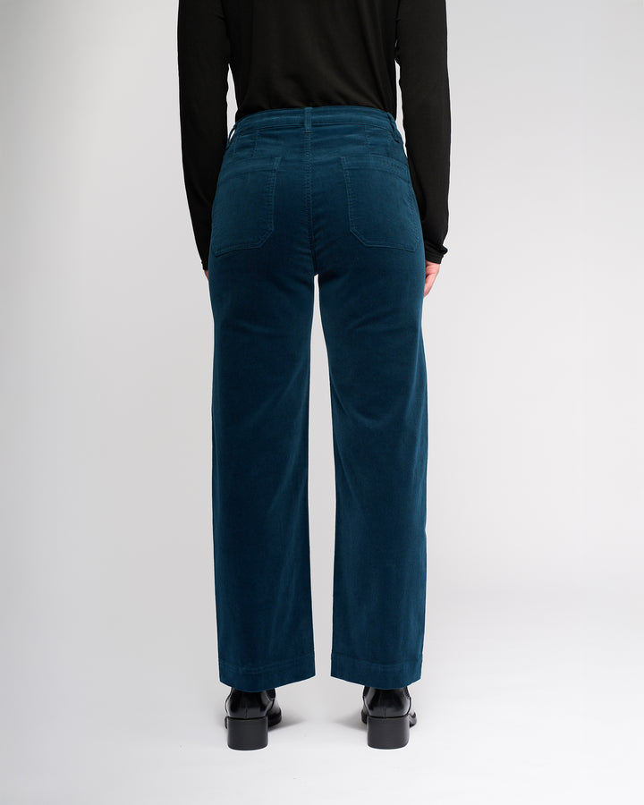 A person stands with their back to the camera, wearing a black long-sleeve top and Vassalli Market Cord Pant in Petrol. The pants feature patch pockets and an ankle-grazer length. The background is plain white.