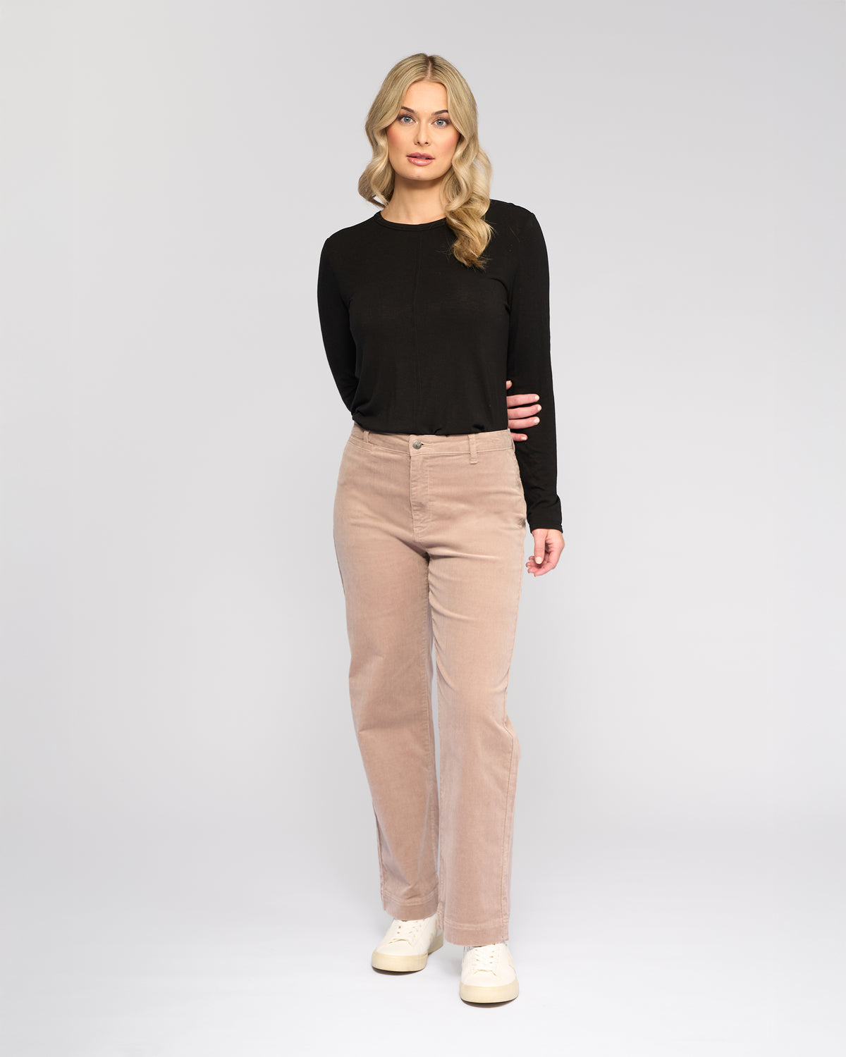 A woman with long blonde hair wears a black long-sleeve shirt, white sneakers, and Vassalli Market Cord Pant - Twig, standing against a plain light gray background with one hand behind her back.