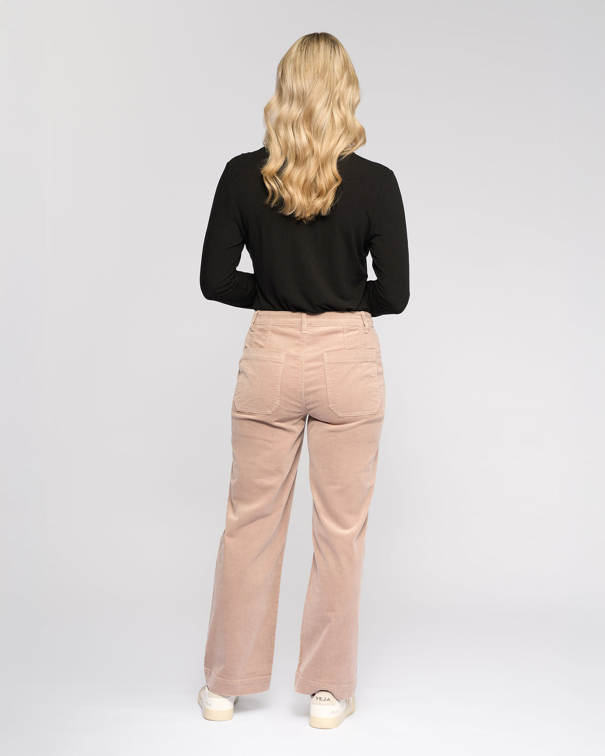 A person with wavy blonde hair in a black long-sleeve top and Vassalli Market Cord Pant - Twig stands facing away against a plain light background, paired with white shoes.
