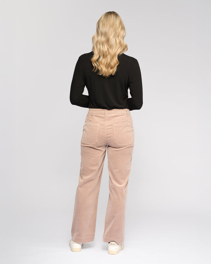 A person with wavy blonde hair in a black long-sleeve top and Vassalli Market Cord Pant - Twig stands facing away against a plain light background, paired with white shoes.