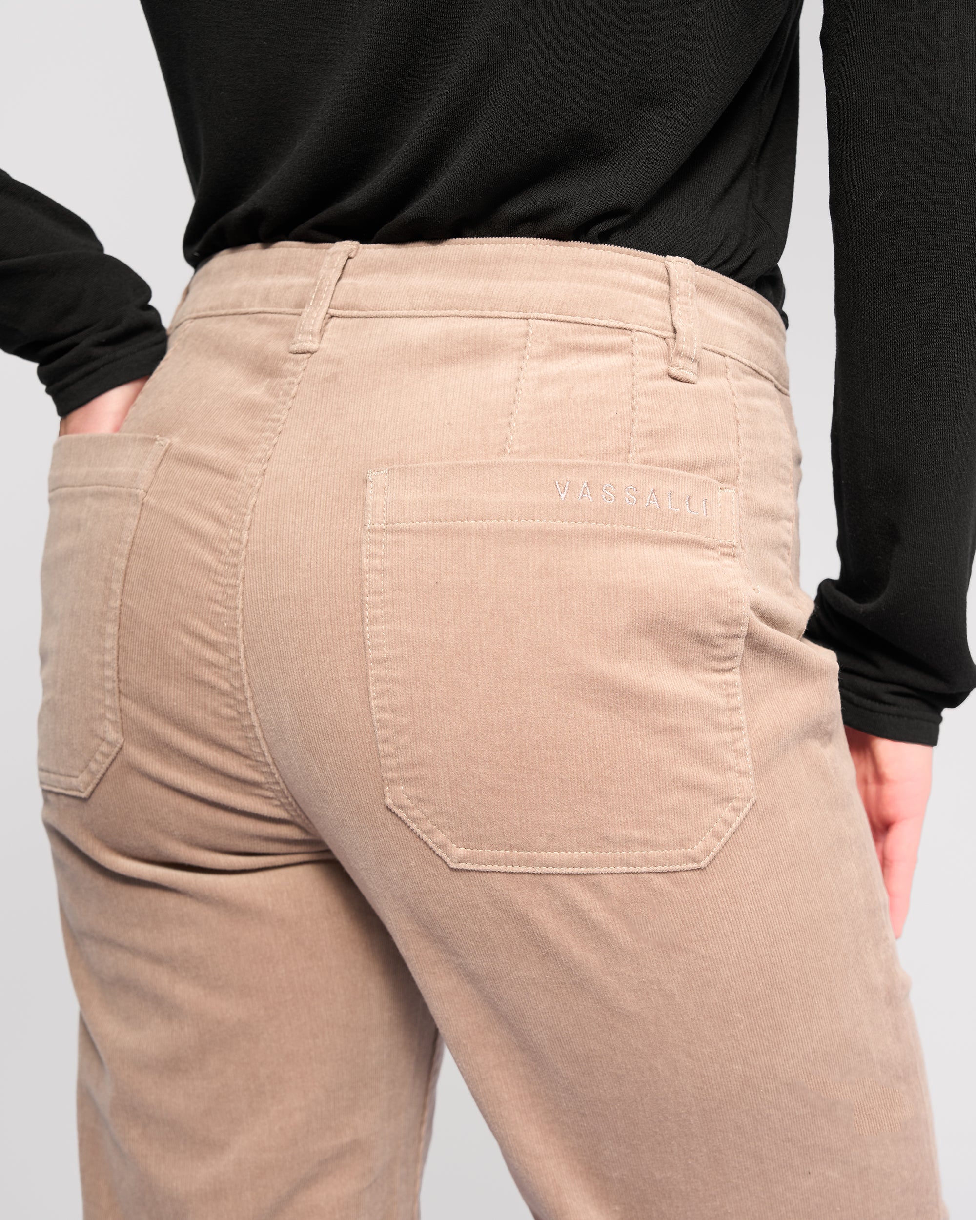 A person is shown from the back wearing Vassalli's Market Cord Pant - Twig, a light beige straight-leg ankle-grazer with two back pockets and "VASSALLI" embroidered above the right pocket, paired with a black top.