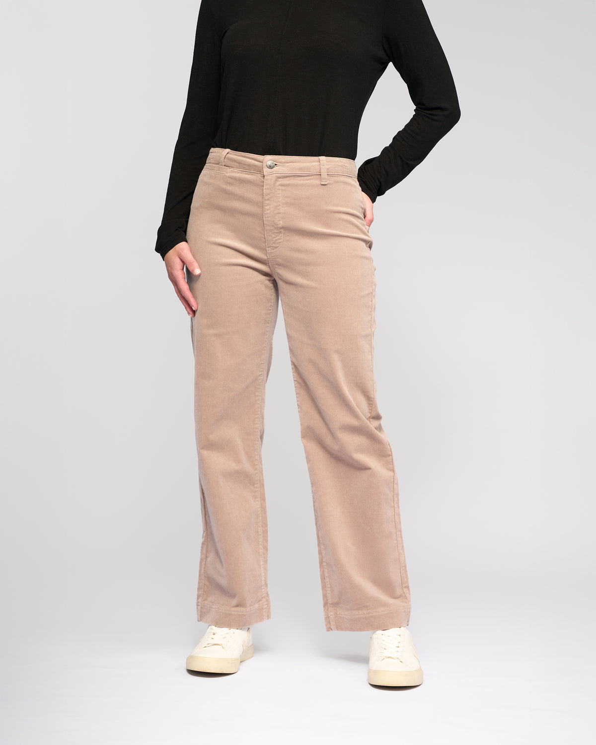 A person wearing a black long-sleeve top, Vassalli Market Cord Pant in Twig, and white sneakers stands against a plain light gray background. The individual's head is not visible in the image.