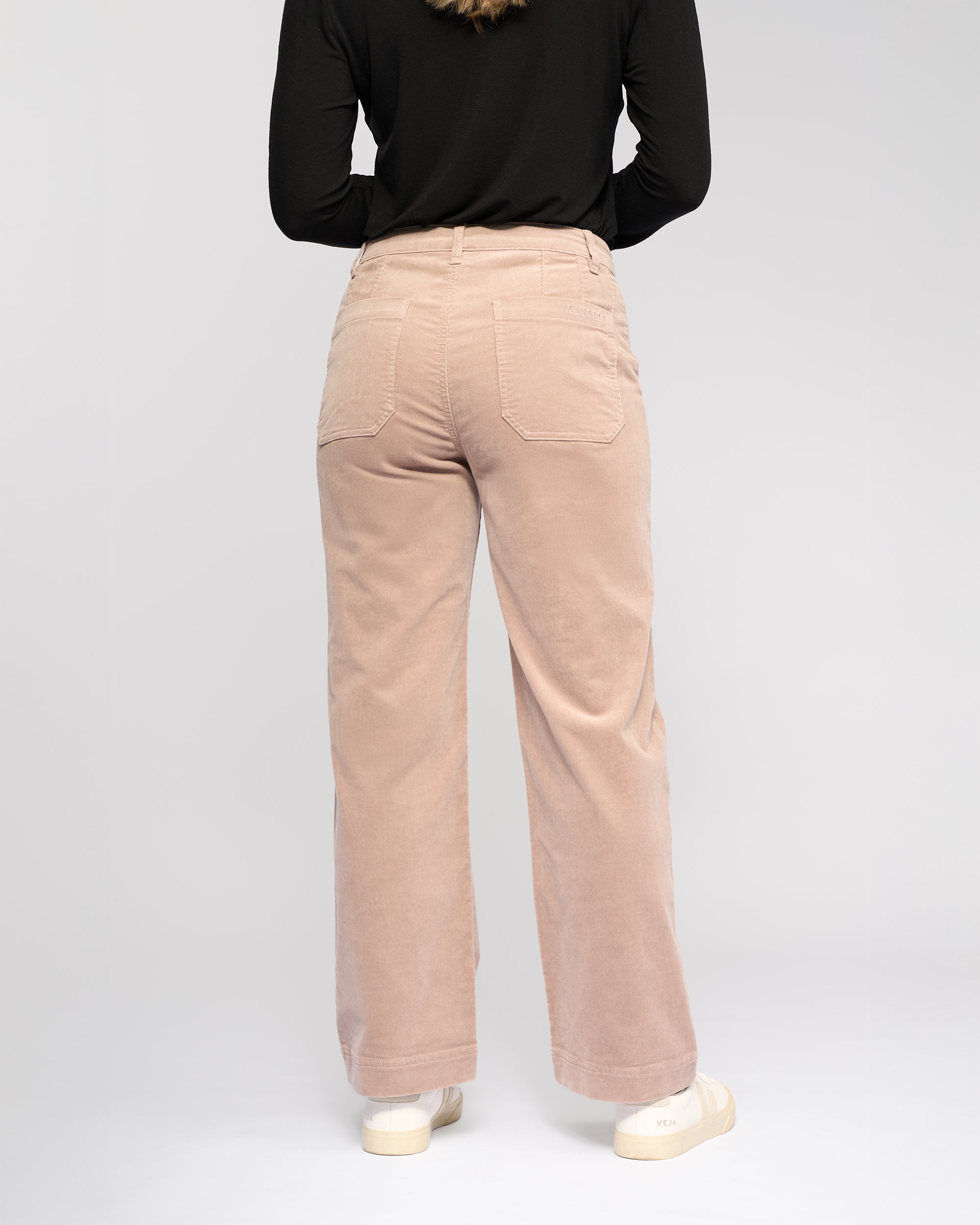 A person stands in a studio wearing Vassalli's Market Cord Pant in Twig, crafted from stretch pinwale cord, paired with a black long-sleeve top and off-white sneakers.