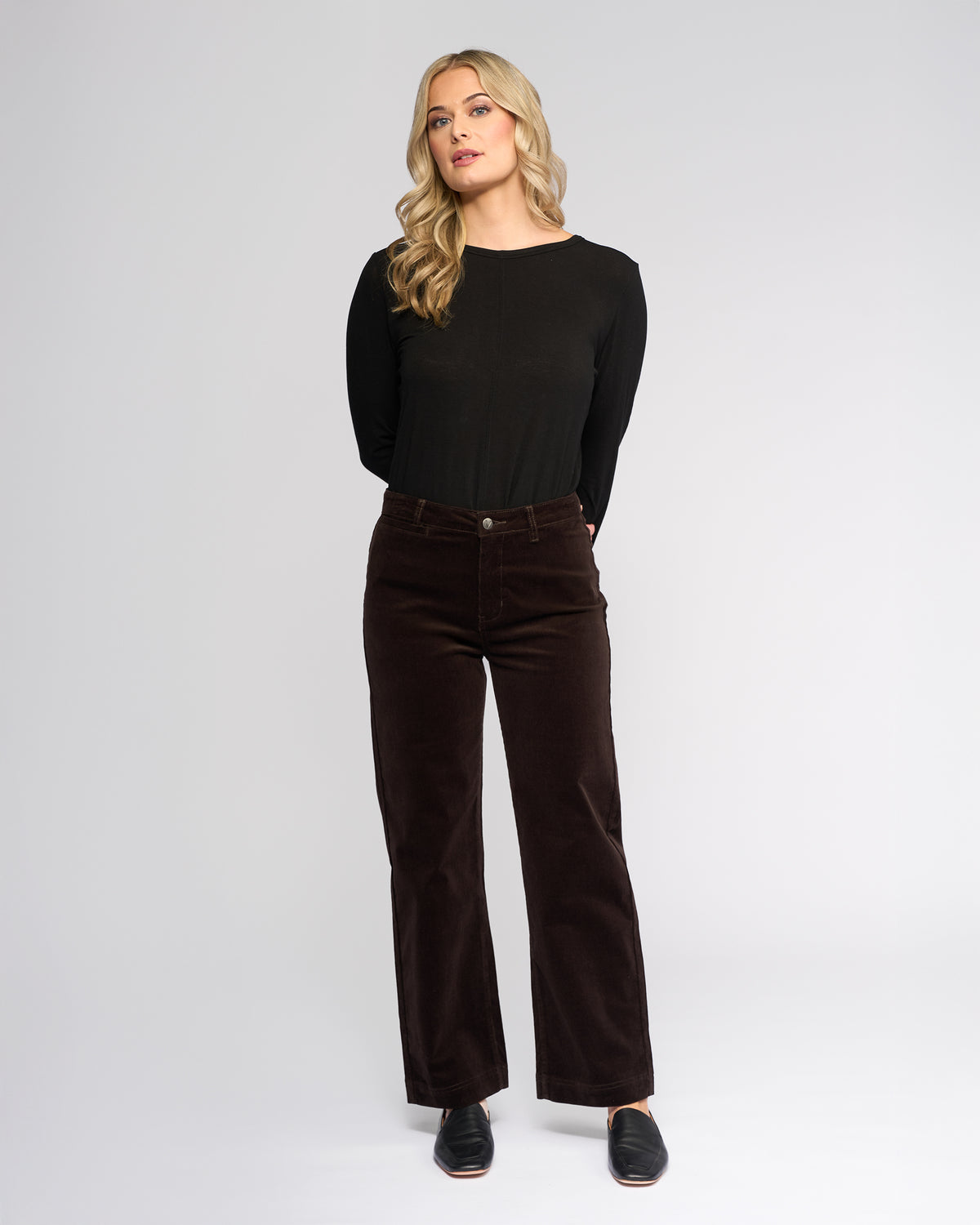 A woman with long blonde hair, wearing a black long-sleeve top, Vassalli Market Cord Pant in Walnut, and black slip-on shoes stands against a plain light gray background with her hands behind her back.