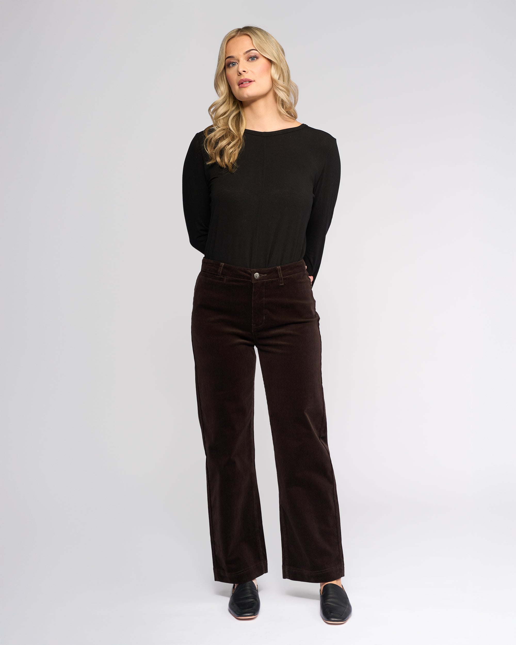 A woman with long blonde hair, wearing a black long-sleeve top, Vassalli Market Cord Pant in Walnut, and black slip-on shoes stands against a plain light gray background with her hands behind her back.