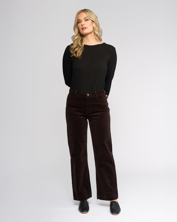A woman with long blonde hair, wearing a black long-sleeve top, Vassalli Market Cord Pant in Walnut, and black slip-on shoes stands against a plain light gray background with her hands behind her back.
