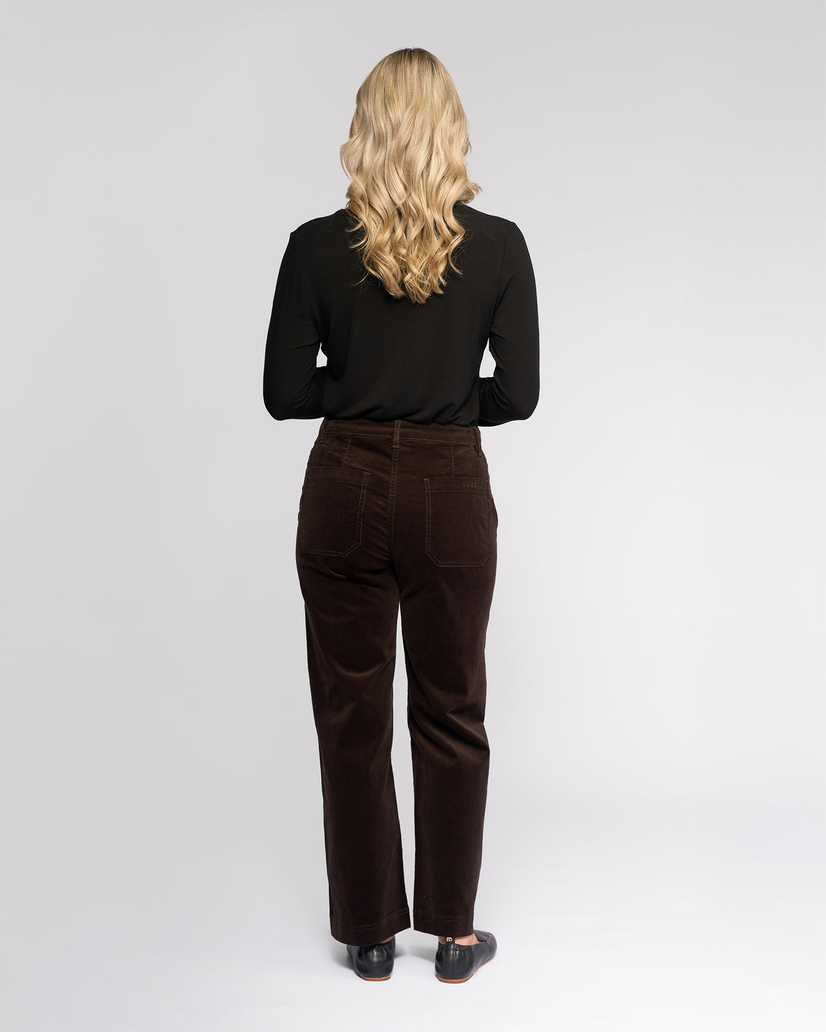 A woman with long, wavy blonde hair faces away from the camera, wearing a black long-sleeve top, black shoes, and Vassalli Market Cord Pant in Walnut against a plain light background.