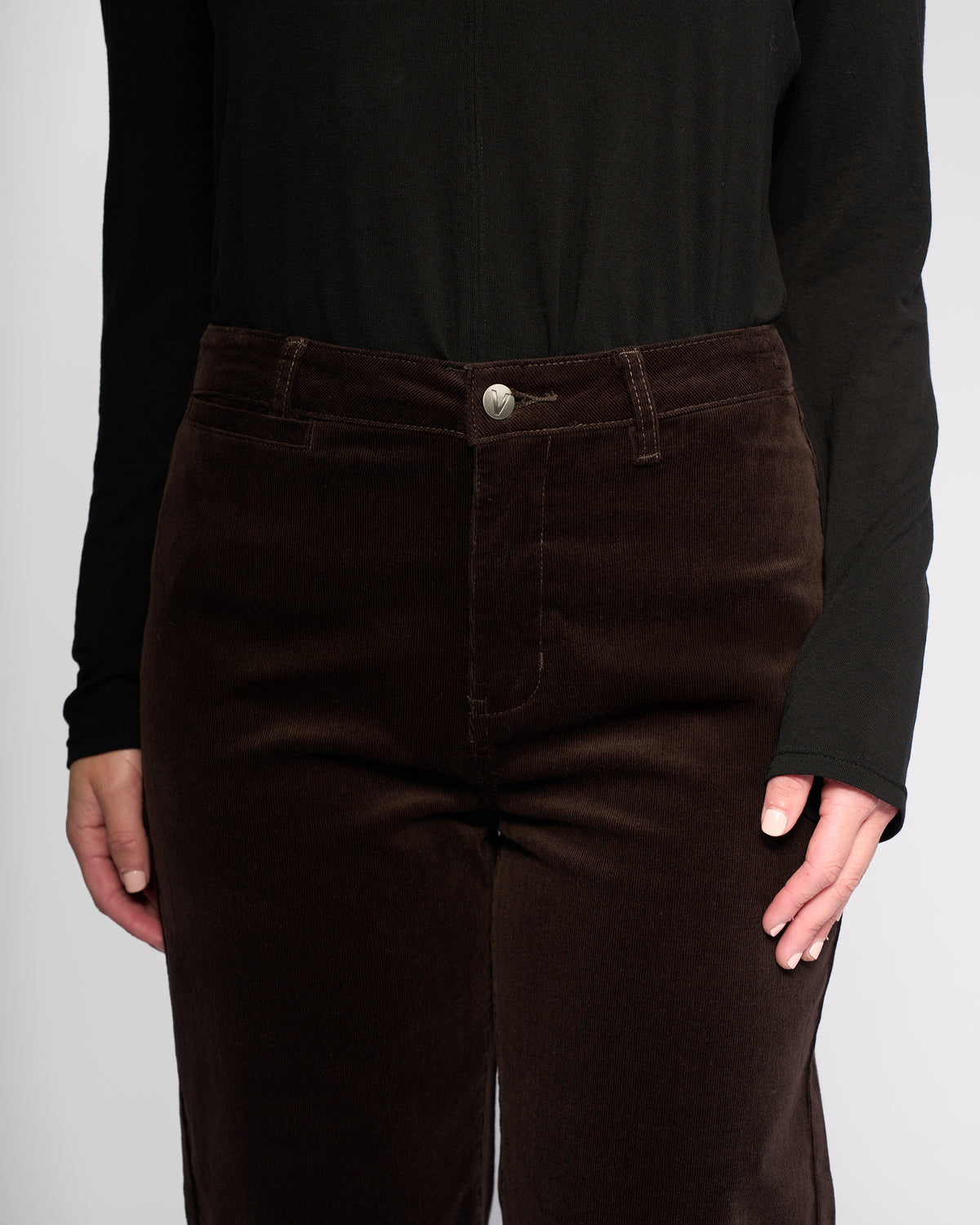 Model in Vassalli’s Market Cord Pant - Walnut, a dark brown straight-leg corduroy style, paired with a black long-sleeve top. Shown from mid-torso to thighs against a plain light background for emphasis on the ankle-grazer fit.