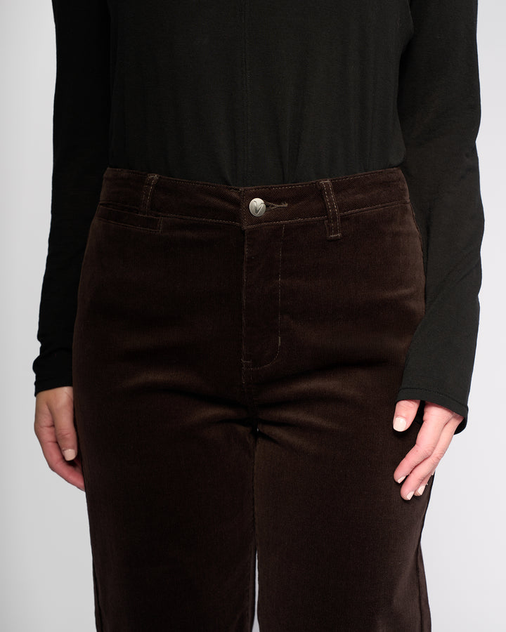 Model in Vassalli’s Market Cord Pant - Walnut, a dark brown straight-leg corduroy style, paired with a black long-sleeve top. Shown from mid-torso to thighs against a plain light background for emphasis on the ankle-grazer fit.