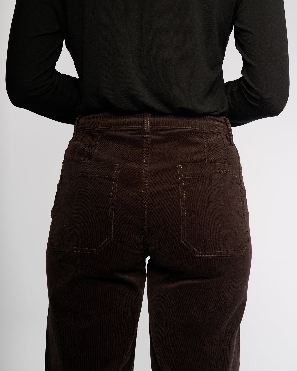 Person stands with their back to the camera, wearing Vassalli’s Market Cord Pant in Walnut and a long-sleeve black shirt. The image highlights the texture and stitching of these straight-leg pinwale cord pants.