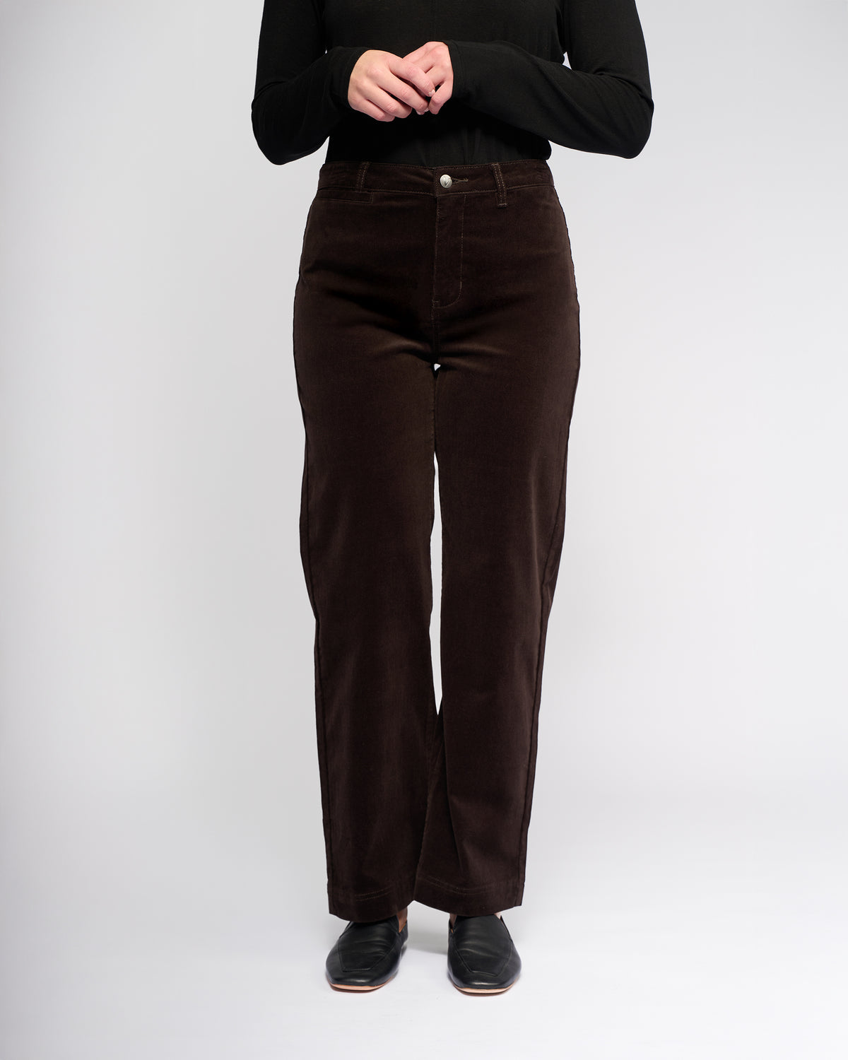 A person stands against a plain background wearing Vassalli’s Market Cord Pant in Walnut, paired with a long-sleeve black shirt and black shoes. Their hands are clasped at chest height, and their face is not visible.