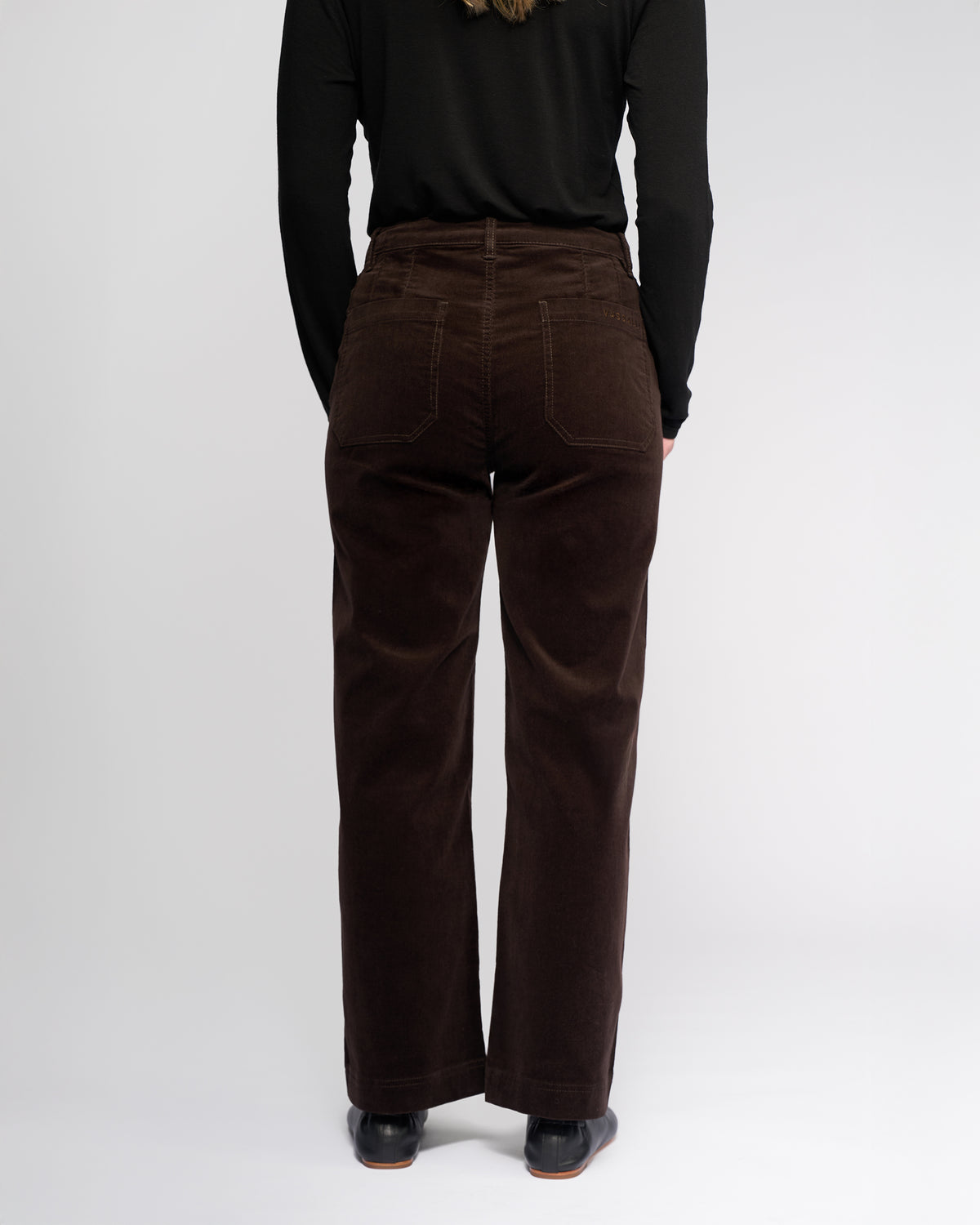 A person models the Vassalli Market Cord Pant in Walnut, paired with a black long-sleeve top, standing with their back to the camera against a white background.