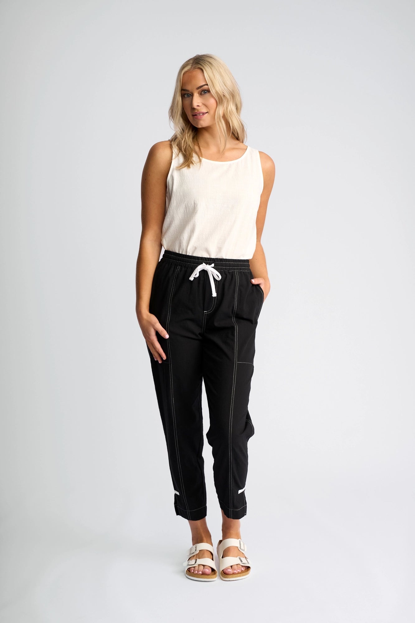 A woman with blonde hair showcases her effortless everyday style wearing the Vassalli Tone Pant - Black, made from 100% cotton and featuring a white drawstring and stitching. She pairs the sleeveless white top with these jogger pants, completes the look with white sandals, and stands sideways with hands in pockets against a plain light background.