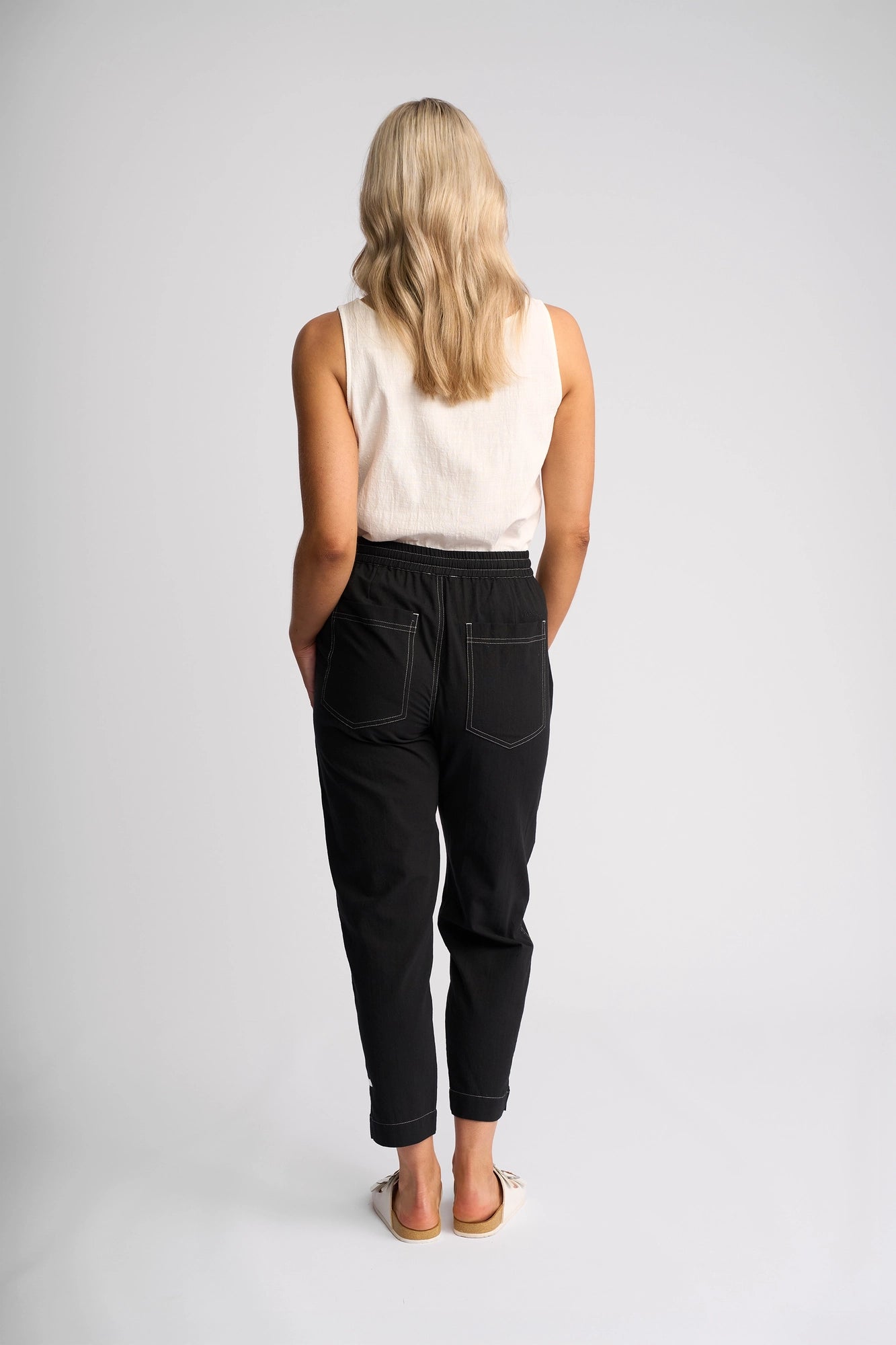 A person with long blonde hair, dressed in a sleeveless white top and Vassalli’s Tone Pant - Black made from 100% cotton, paired with sandals, stands facing away from the camera against a plain white background—exemplifying effortless everyday style.