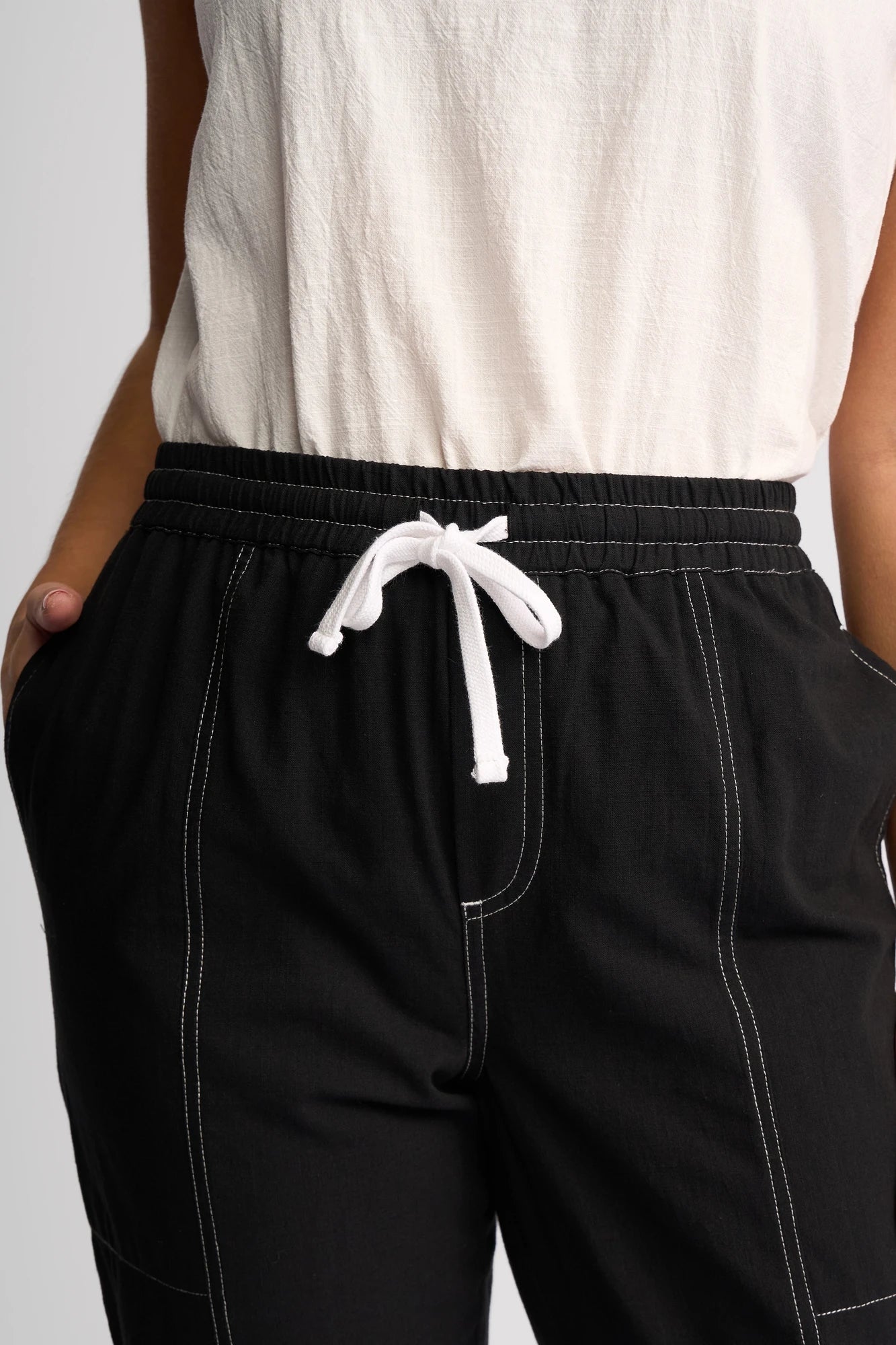 A person wears the Vassalli Tone Pant - Black, featuring distinctive white stitching, paired with a white shirt and hands in pockets. Made from 100% cotton for comfort and breathability, this look embodies effortless everyday style. The image is cropped to showcase only the lower torso and upper legs.
